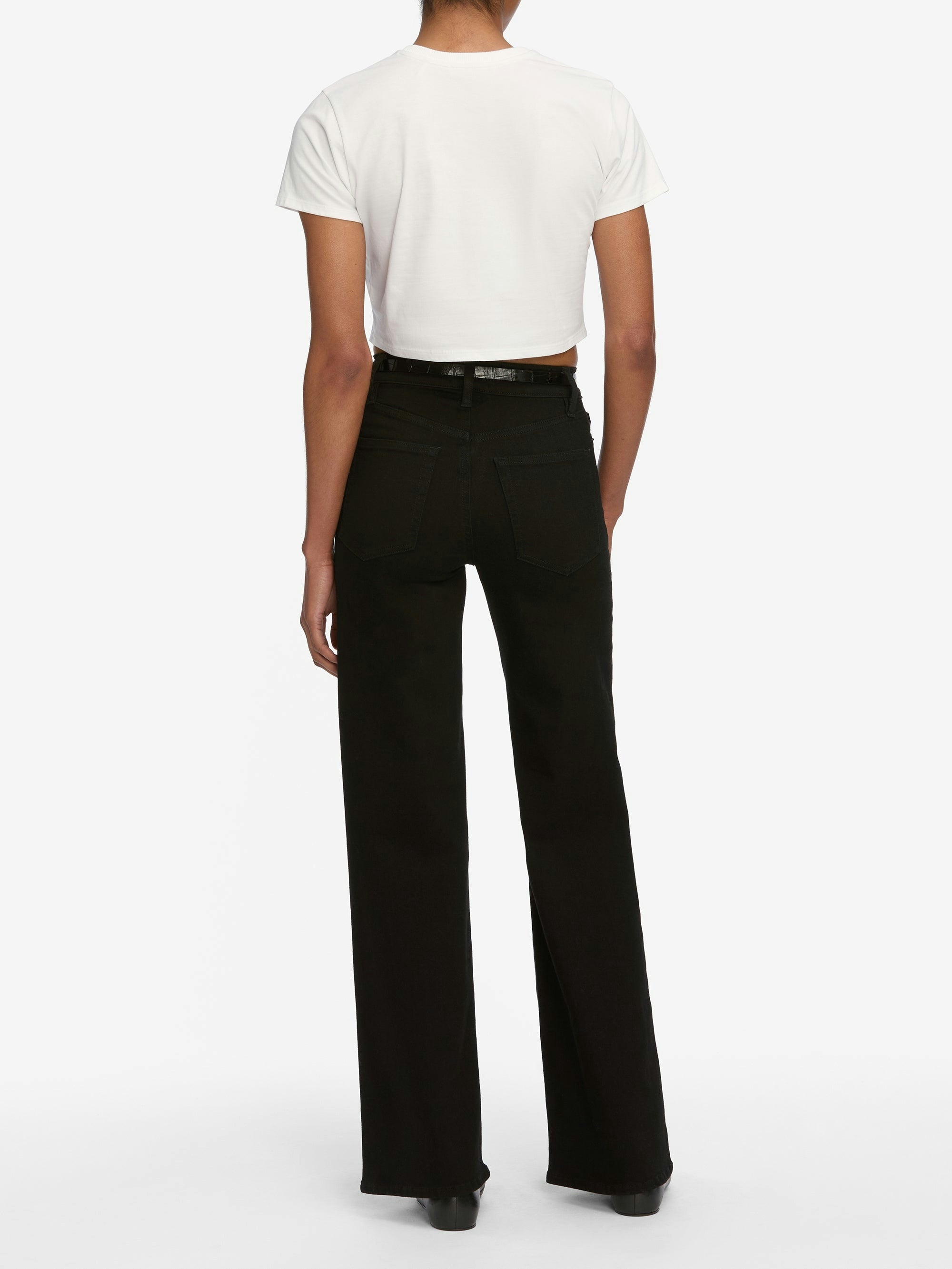 A person stands facing away, wearing a white cropped T-shirt, black Frame Le Slim Palazzo Jeans by Frame, and black shoes against a plain white background.