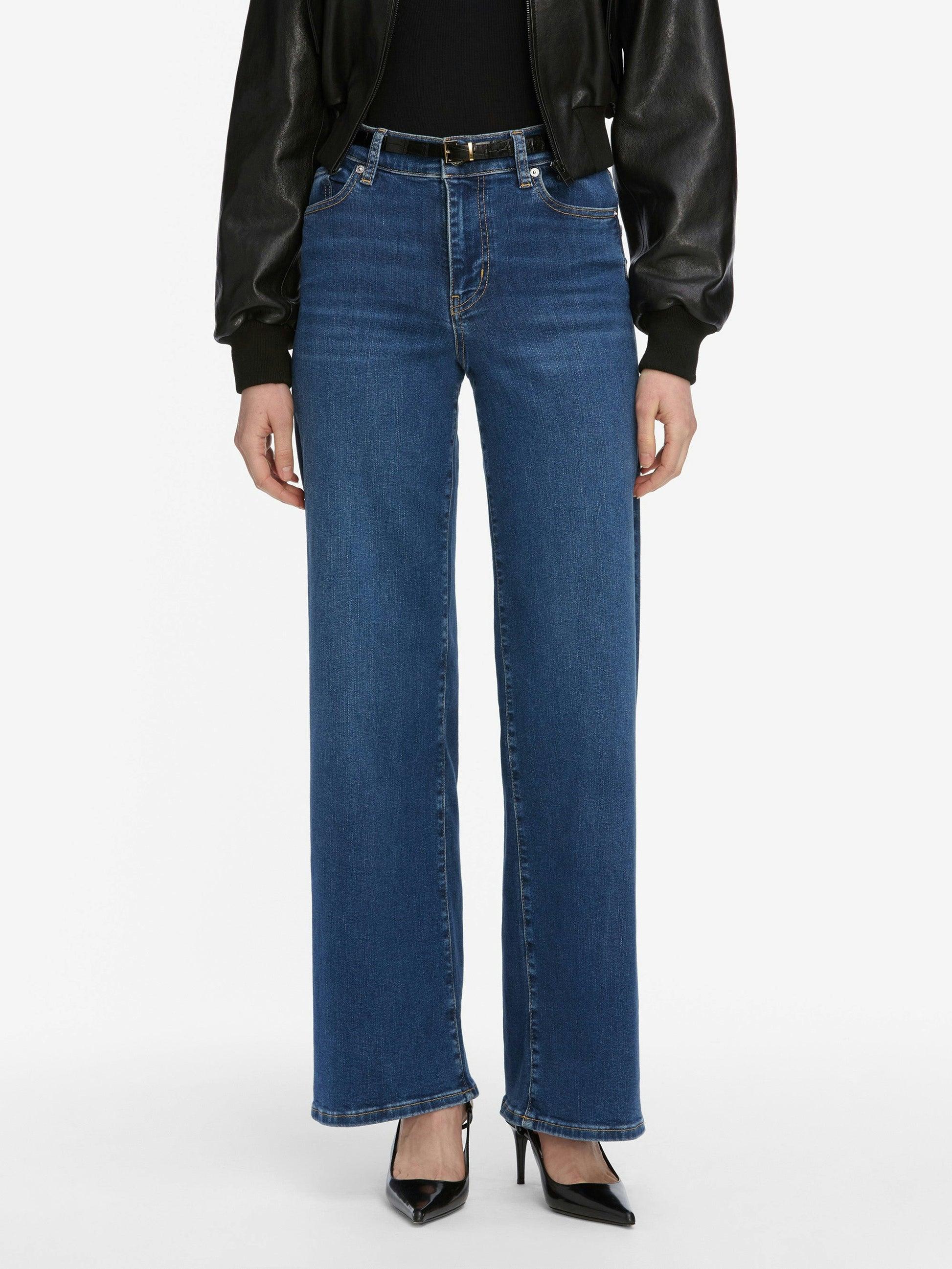 A person wears Frame Le Slim Palazzo Jeans - Femma by Frame, styled with a black belt, black leather jacket, and black pointed-toe heels, standing against a plain white background.