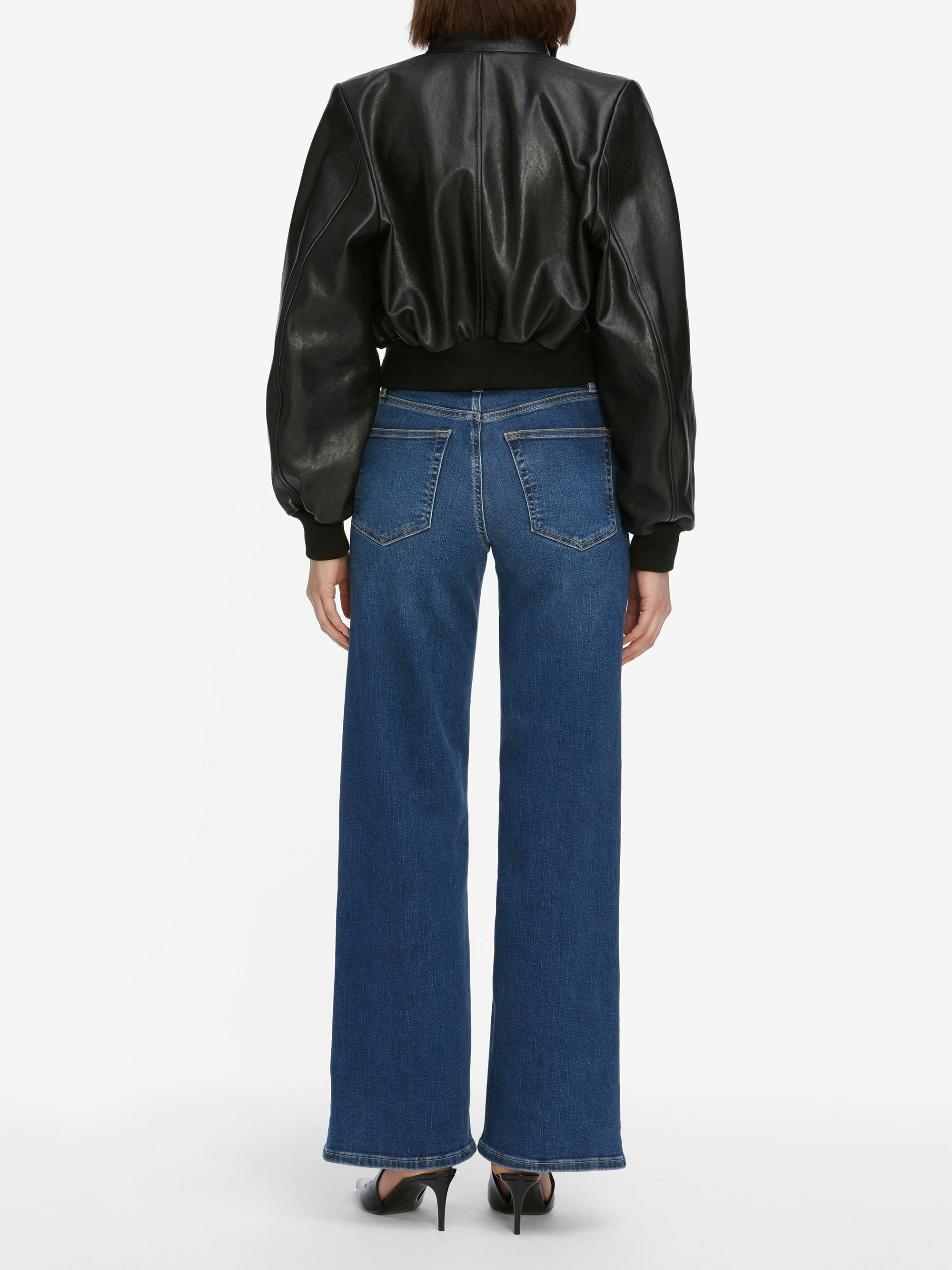 A person stands facing away in a black leather jacket, Frame Le Slim Palazzo Jeans - Femma by Frame, and black heels against a plain white background.