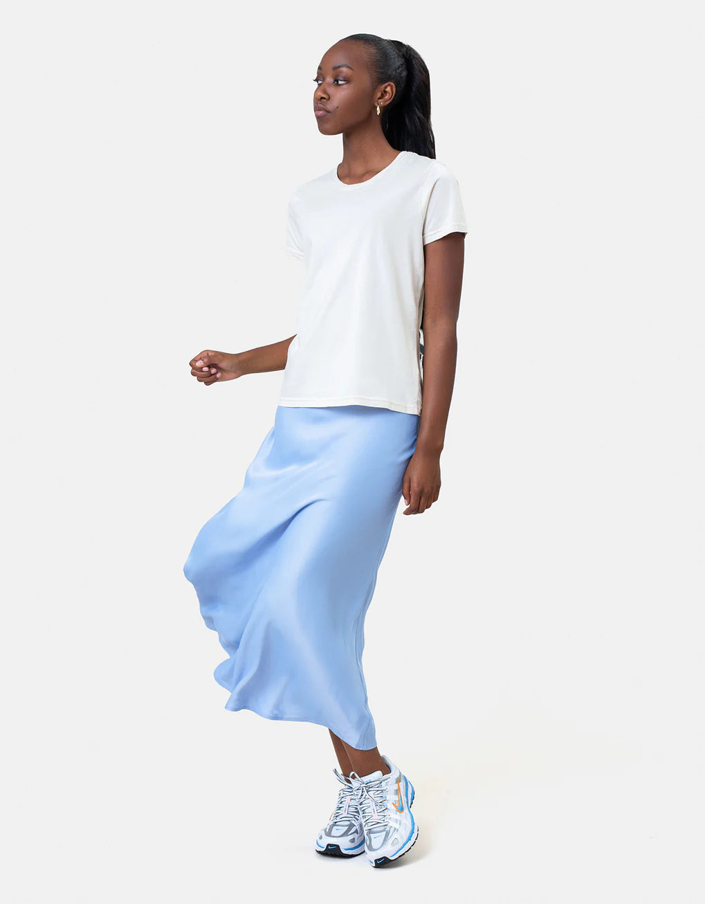 A woman in a white Light Organic Tee from Colorful Standard and a blue skirt paired with sneakers walks to showcase an outfit.