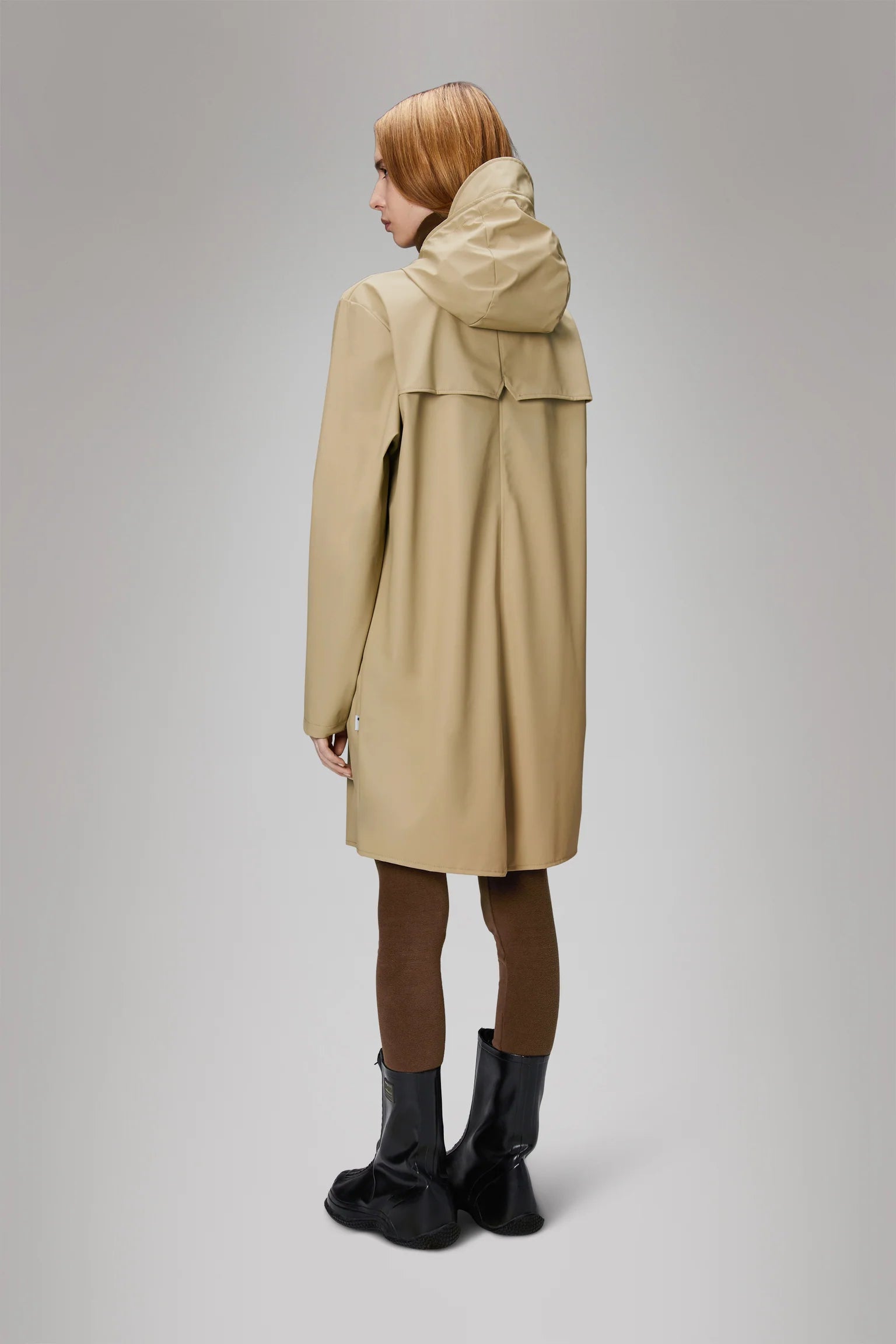 Person with shoulder-length hair facing away, wearing a hooded beige Long Jacket from Rains' Core Colours collection, made from Rains' signature PU fabric, paired with brown leggings and black boots against a plain background.