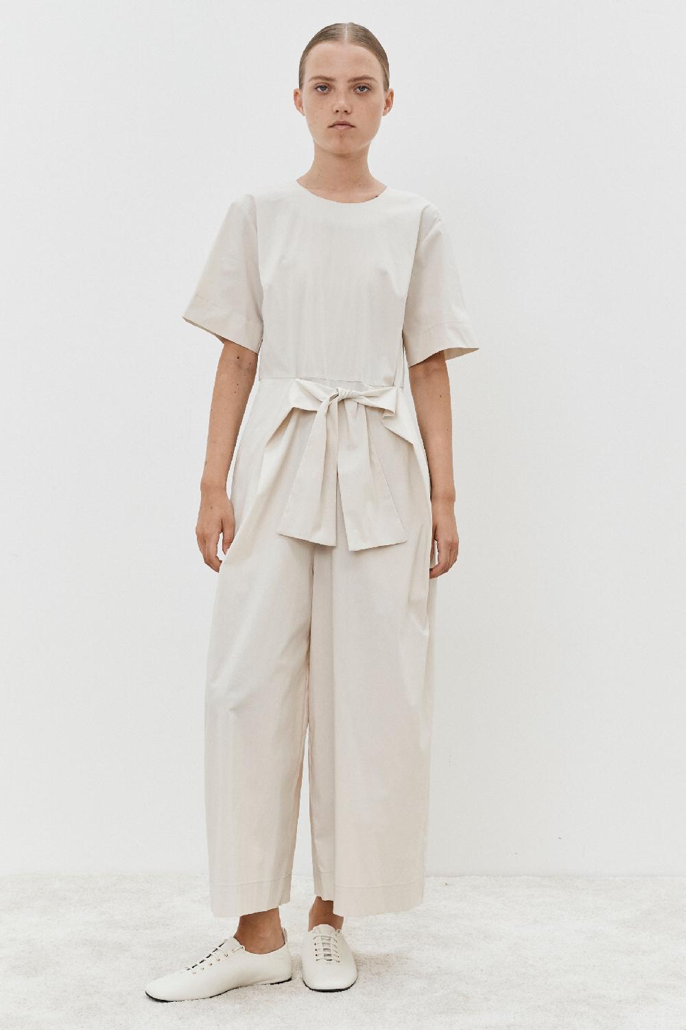 A person stands against a plain white background in the Jumpsuit - Oyster by MeiMeij, crafted from a luxurious cotton blend with a waist tie, and paired with white shoes.