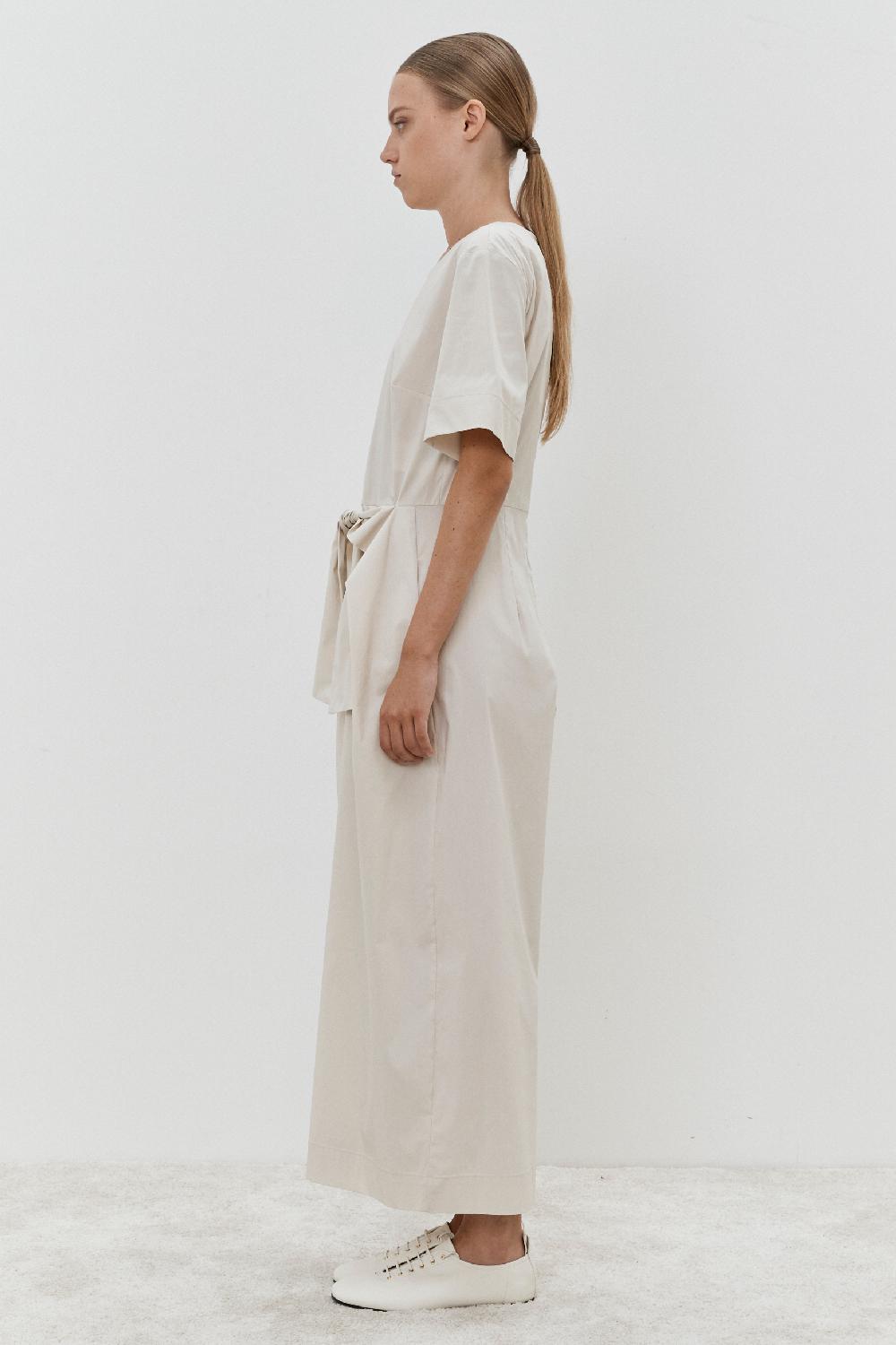A person with long hair tied back wears an ankle-length, light-colored MeiMeij Jumpsuit - Oyster with short sleeves and pairs it with white shoes, standing in profile against a plain white background.