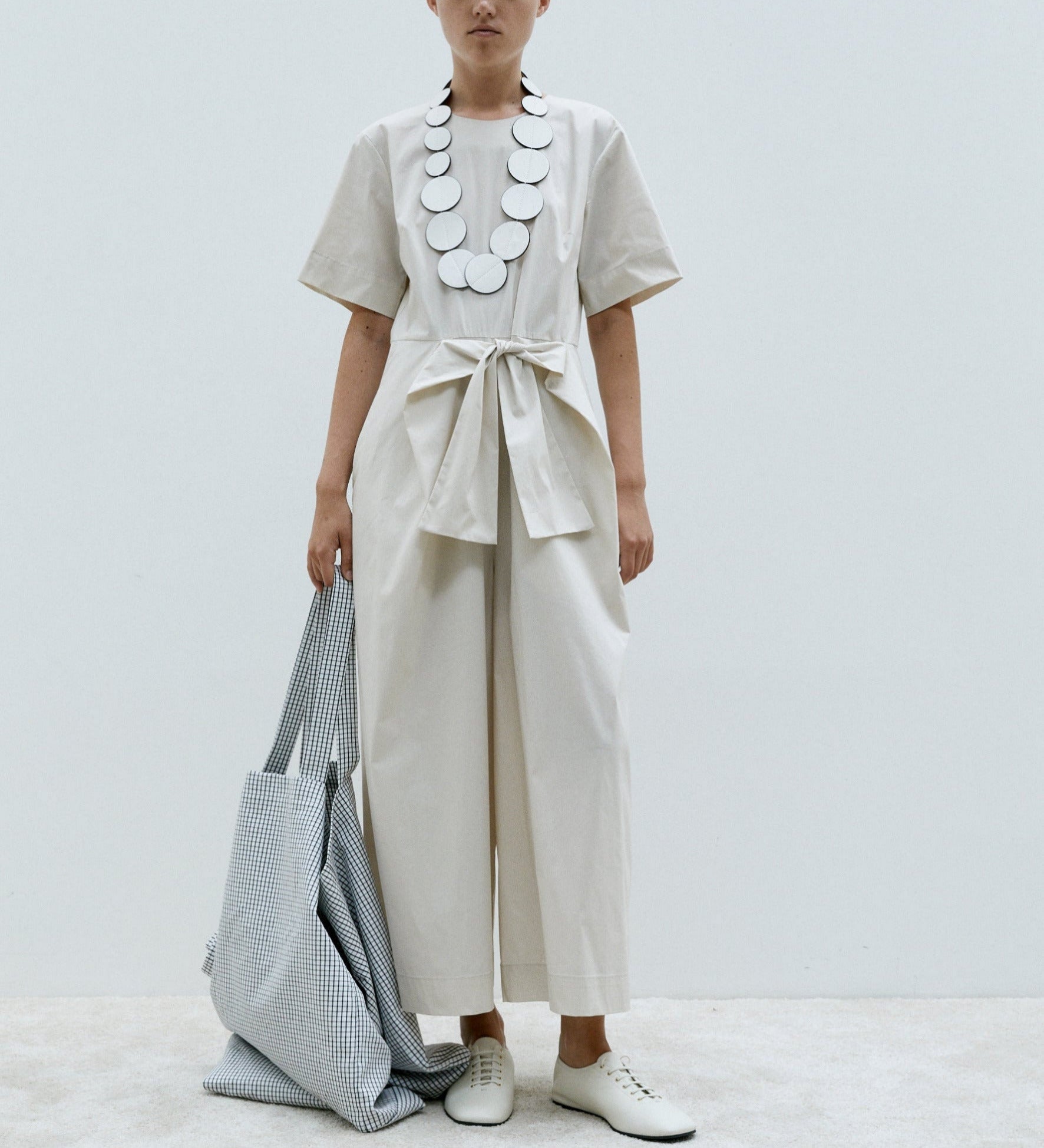 a woman wears a light coloured jumpsuit by meimeij with a tie belt and short sleeves, and white shoes and a silver necklace and a gingham bag