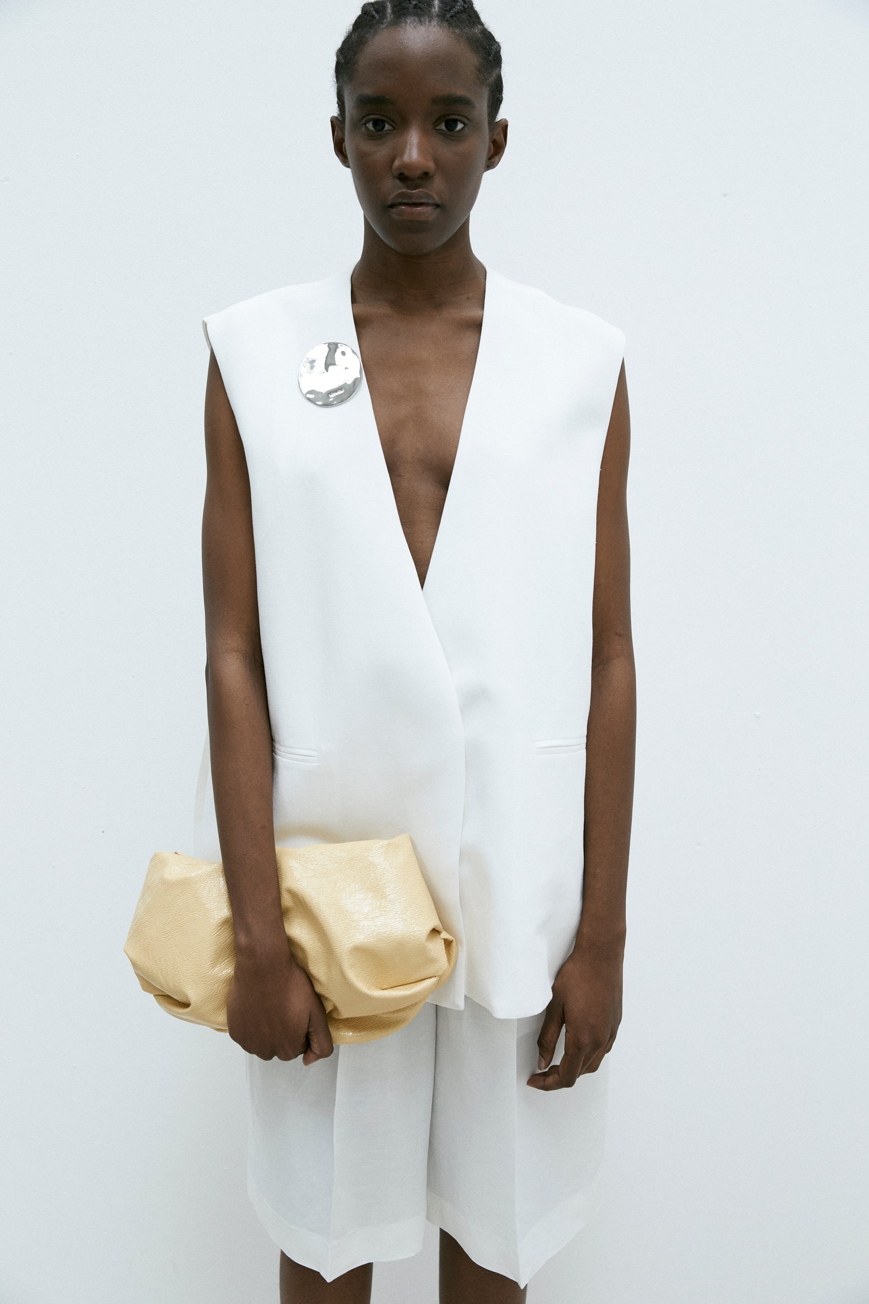 A person wearing MeiMeij's Oversized Vest in Off White and shorts, holding a gold clutch bag. This ensemble showcases a modern fit that perfectly balances effortless style and comfort, ideal for those who appreciate the elegance of Italian sizing.
