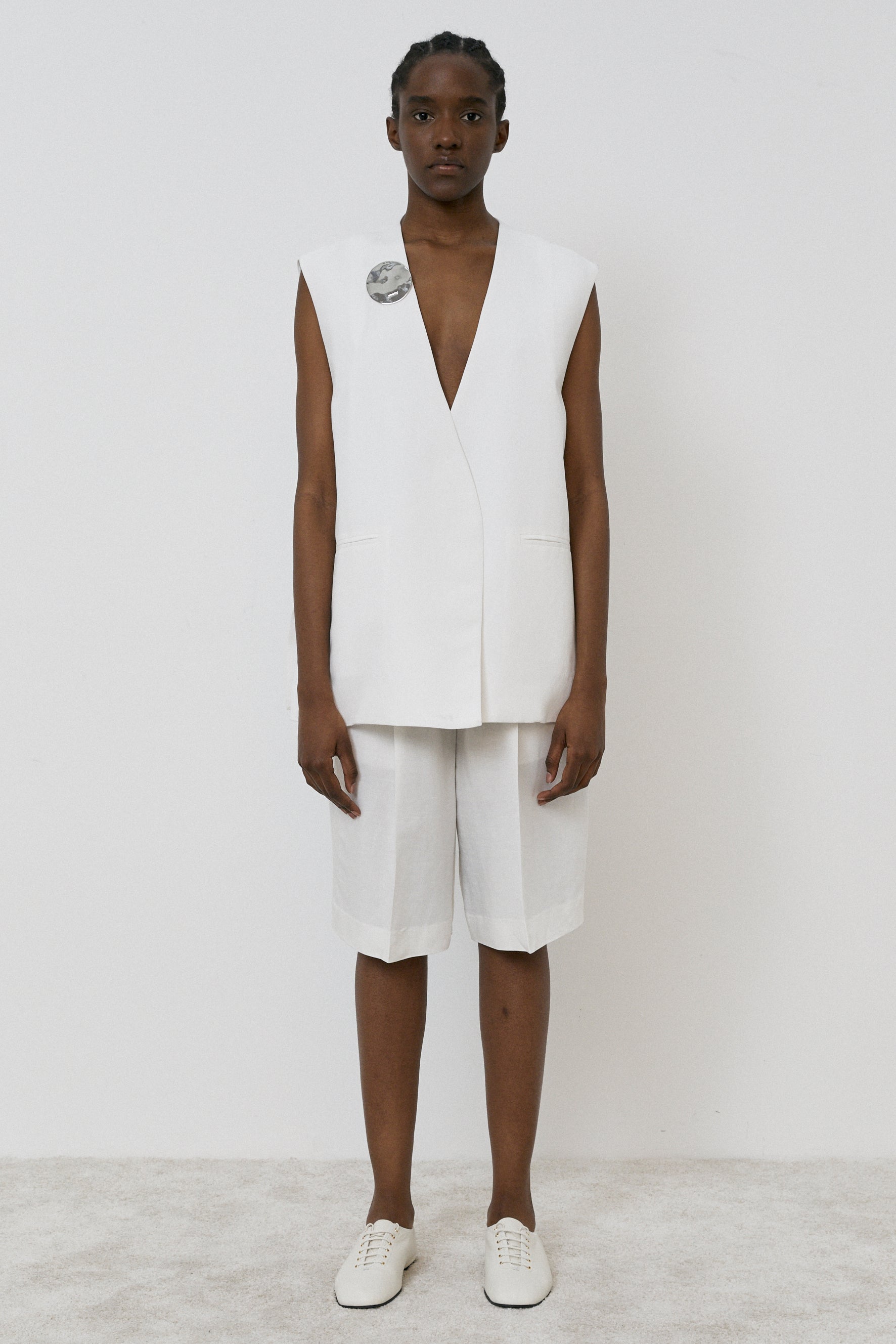 A person poses on a light floor against a white backdrop, wearing MeiMeij's Oversized Vest in Off White paired with knee-length shorts and sleek white shoes. The ensemble’s modern fit and elegant Italian sizing are enhanced by a round pin for added flair.