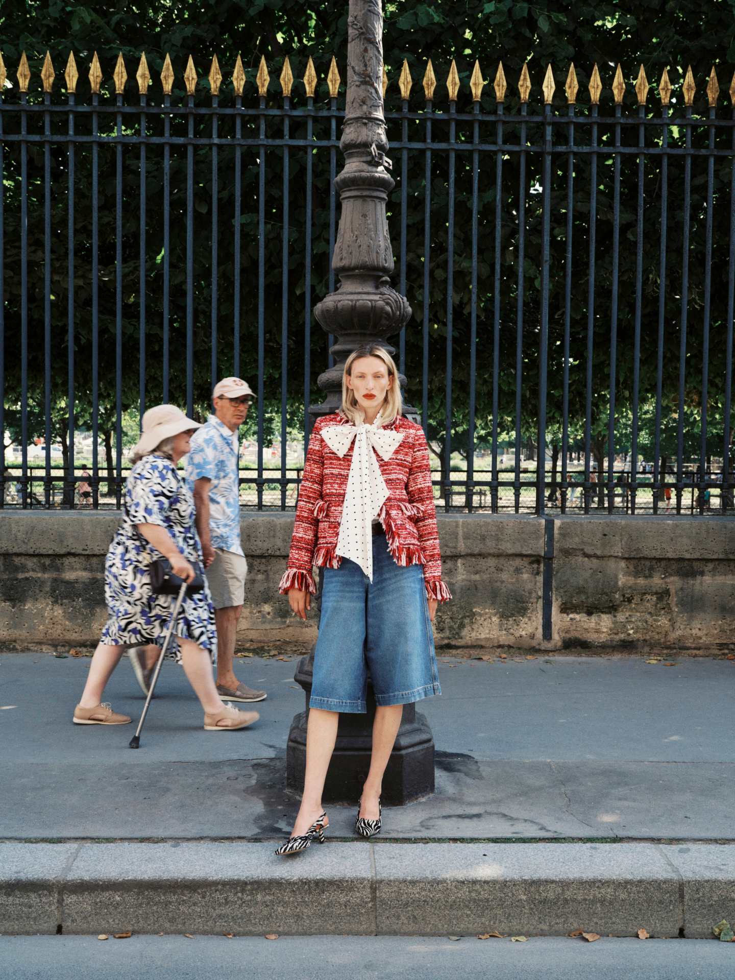 Wearing the Essentiel Antwerp Iskra Bouclé Jacket in burgundy red, a woman stands by a lamp post in a boxy silhouette; behind her, two people walk along the sidewalk.