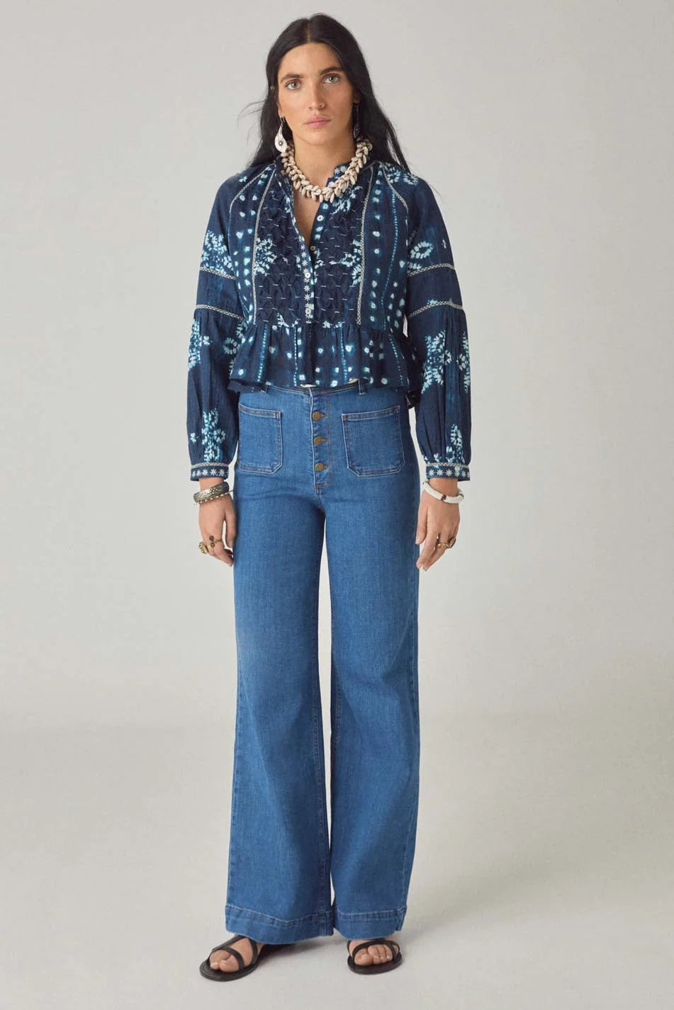 A woman faces forward against a plain light background, wearing the Maison Hotel Sora Blouse in Ocean Blue, paired with high-waisted button-front jeans, black sandals, and shell jewelry.
