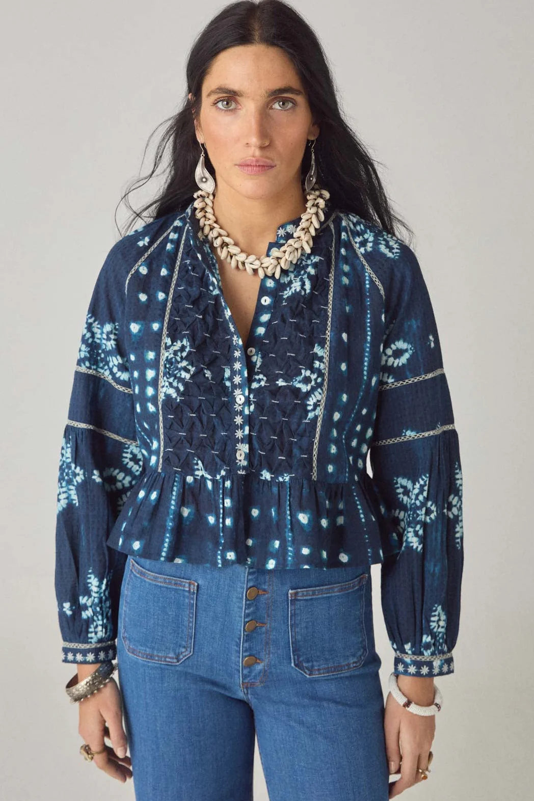 A woman with long dark hair wears the Maison Hotel Sora Blouse in Ocean Blue, featuring ruffle details and a peplum hem, paired with high-waisted blue jeans and shell and beaded jewelry, against a plain background.