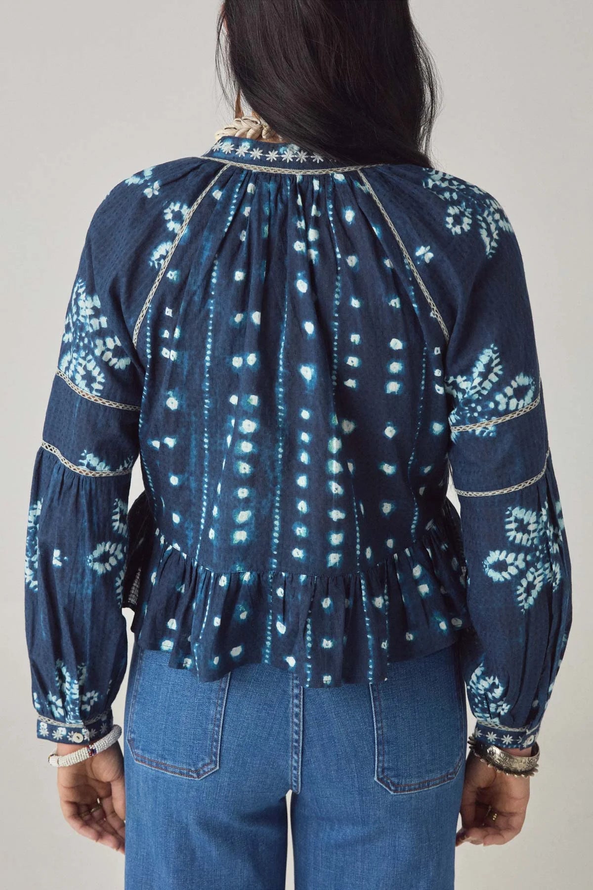 A person shown from the back wears the Maison Hotel Sora Blouse in Ocean Blue, featuring a dark blue and white tie-dye print with a peplum hem, paired with blue jeans.