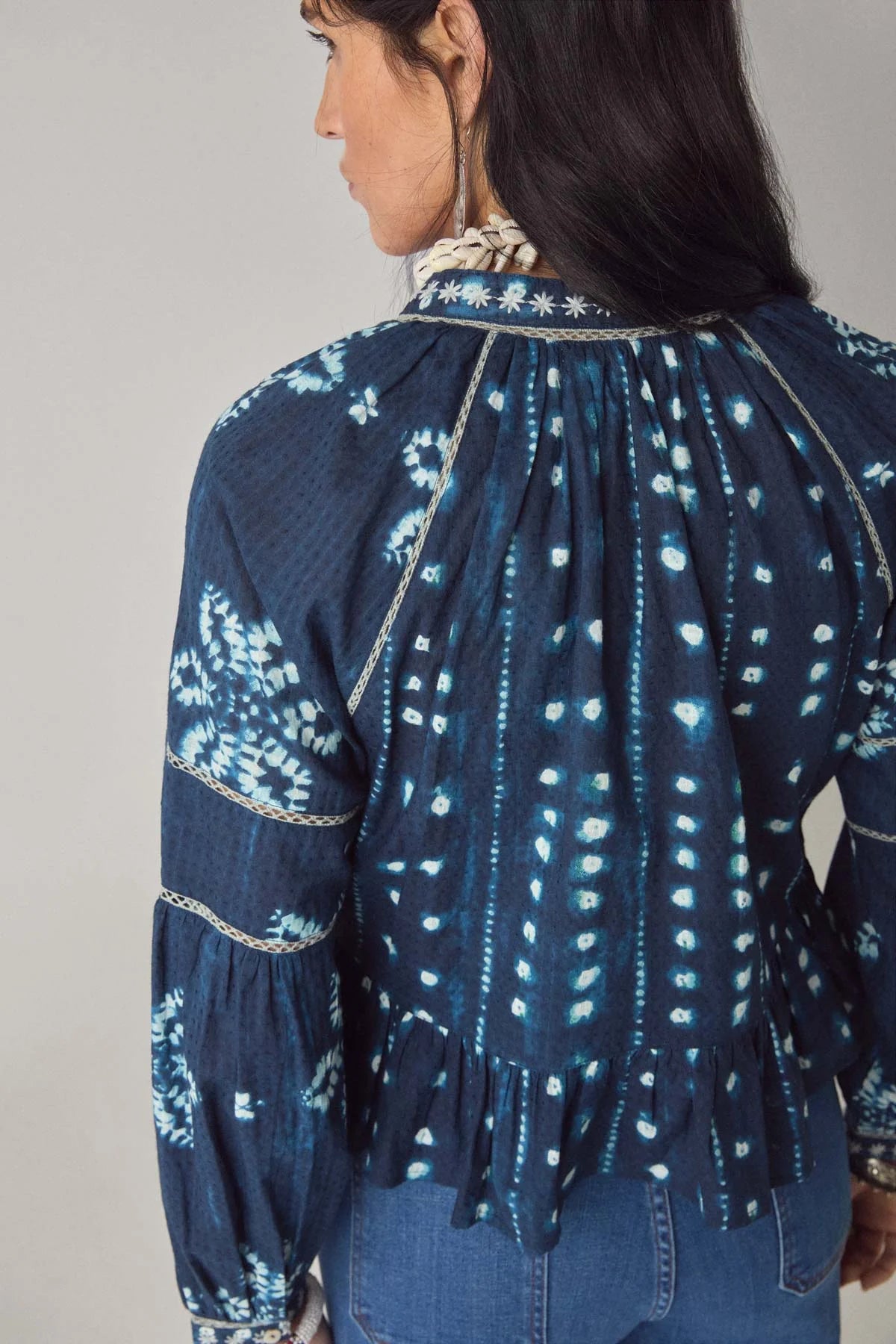 A woman is seen from the back wearing the Maison Hotel Sora Blouse in Ocean Blue, featuring long sleeves, a white tie-dye print, and a ruffled peplum hem.