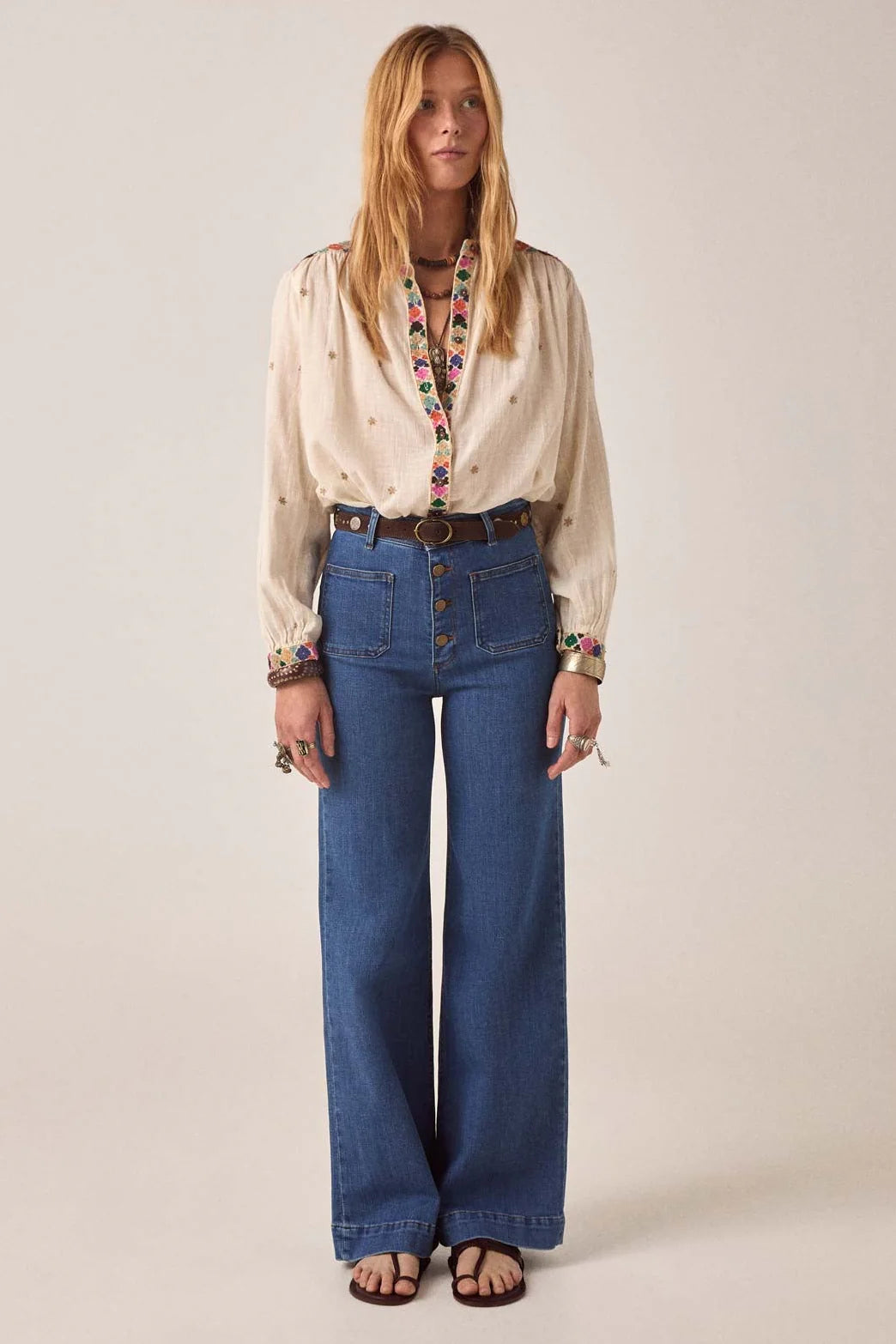 A person stands facing forward wearing the Maison Hotel Cleo Blouse in Off White, high-waisted wide-leg blue jeans, a brown belt, and black sandals against a plain background.