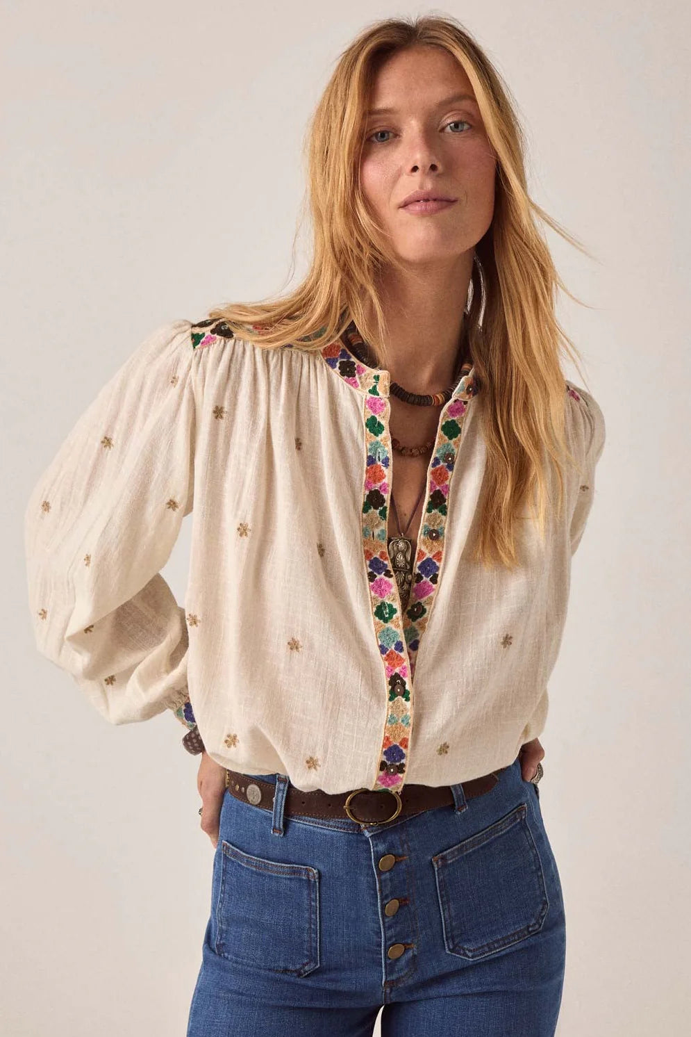 A woman wearing the Maison Hotel Cleo Blouse in Off White, featuring gold embroidery and floral trim, pairs it with high-waisted blue jeans and stands with hands in pockets against a plain background.