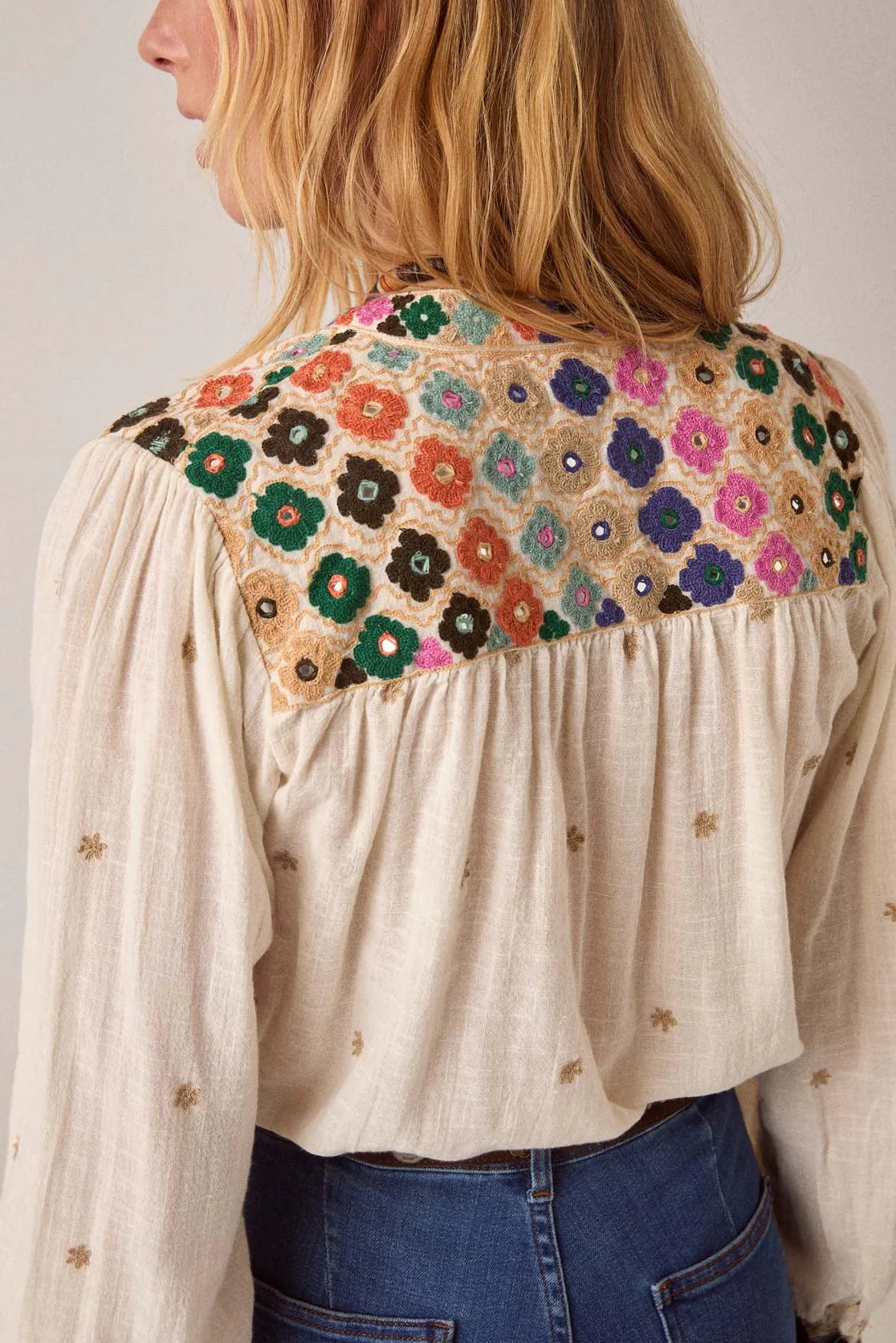 A person wearing the Cleo Blouse - Off White by Maison Hotel, featuring delicate gold embroidery and a colorful floral yoke, paired with blue jeans.
