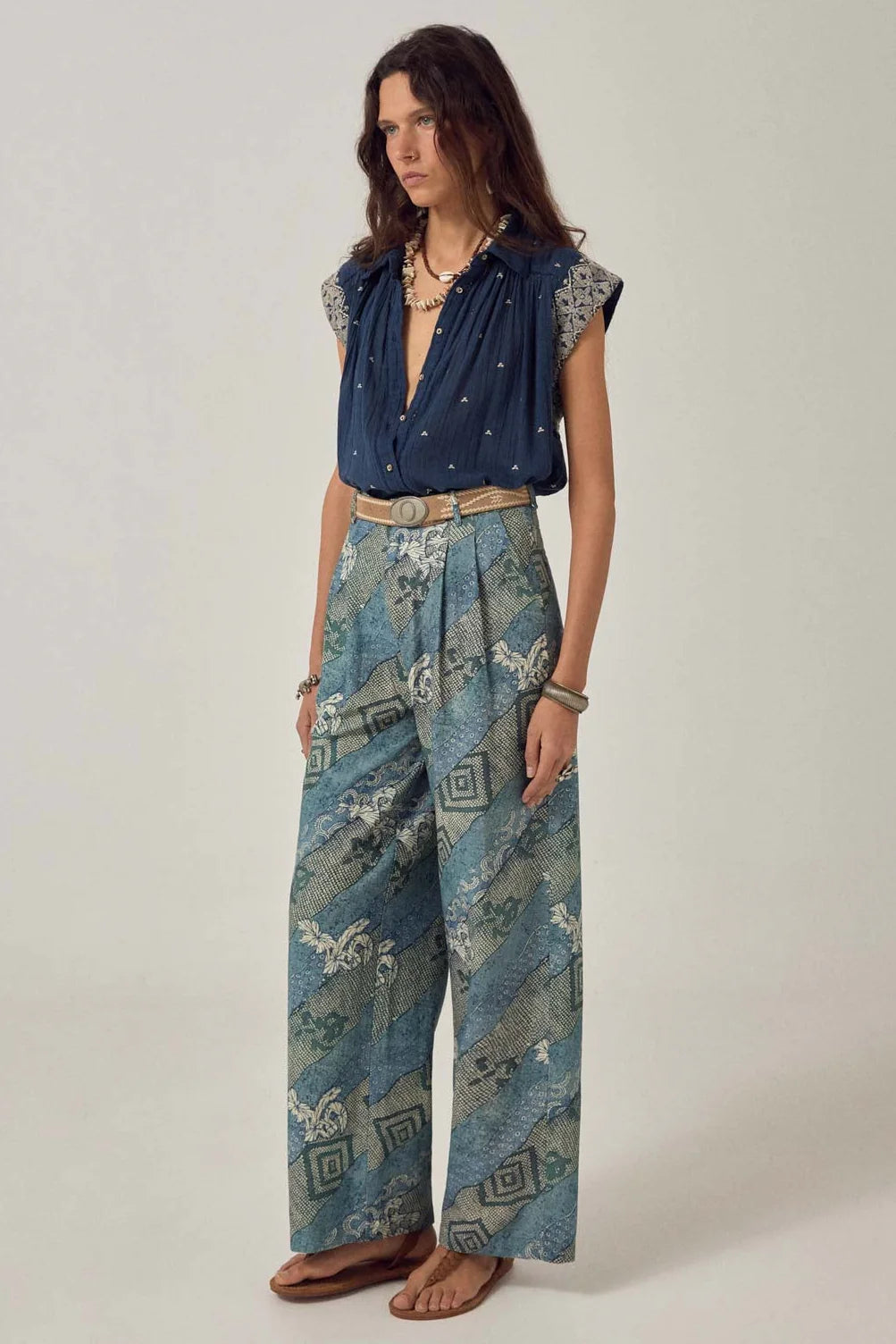 Wearing the Maison Hotel Karina Sleeveless Shirt in Deep Navy, wide-leg blue patterned pants, a beige belt, sandals, and layered jewelry with a lightweight cotton vest, the individual stands against a plain background.