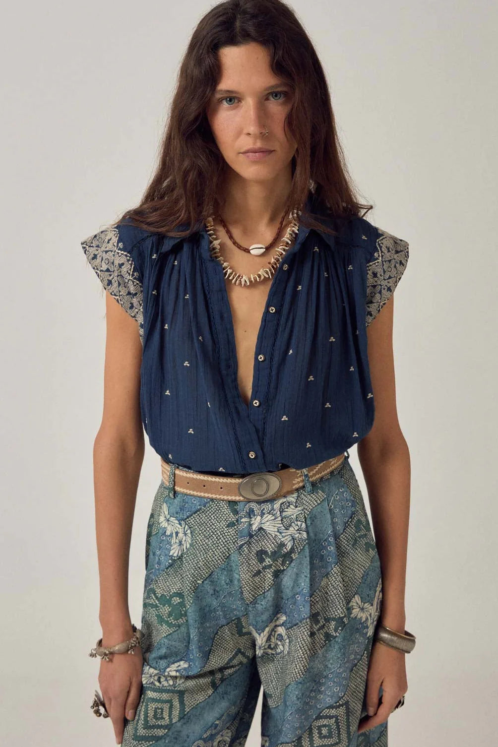 Woman with long brown hair wears the Maison Hotel Karina Sleeveless Shirt in Deep Navy, paired with patterned blue-green trousers, a beige belt, shell necklace, silver bracelets, and a cotton vest against a plain background.