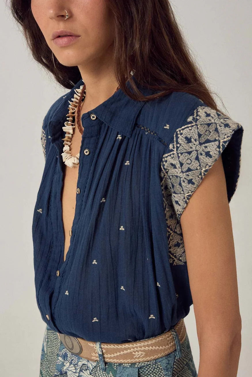 A woman wears the Maison Hotel Karina Sleeveless Shirt in Deep Navy, styled with a shell necklace, patterned belt and skirt, and a lightweight cotton vest for added texture.