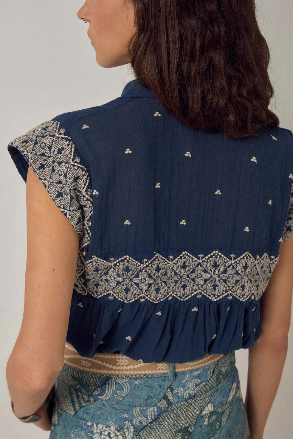 A person with wavy brown hair is seen from the back wearing the Maison Hotel Karina Sleeveless Shirt in Deep Navy, a lightweight cotton top embroidered with white patterns.