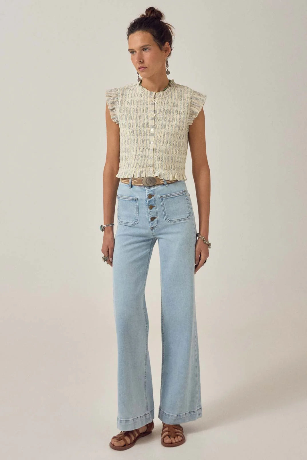 A woman stands against a plain background wearing the Maison Hotel Omaris Sleeveless Blouse in light blue, paired with high-waisted flare jeans and brown sandals.