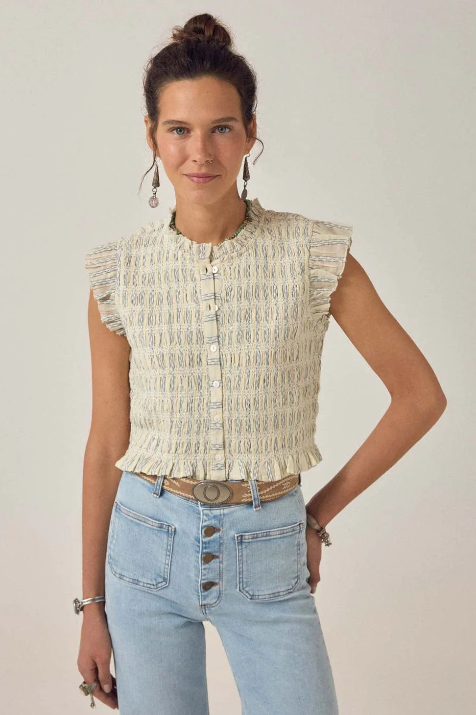 A woman faces forward wearing the Omaris Sleeveless Blouse in light blue by Maison Hotel, paired with high-waisted jeans, earrings, and bracelets. Her hair is styled in a bun.