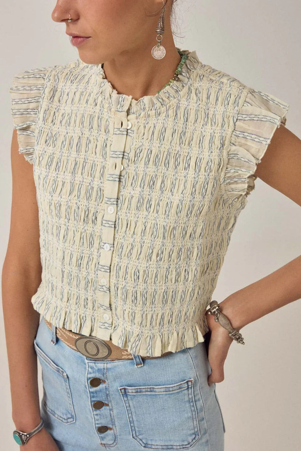 A person in the Maison Hotel Omaris Sleeveless Blouse - Light Blue paired with high-waisted jeans and assorted jewelry, standing with one hand on their hip.