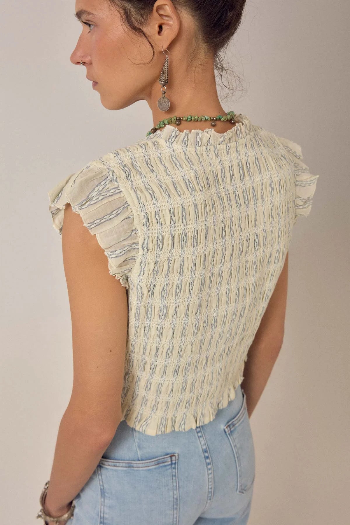 A person is shown from the back in a neutral setting wearing the Omaris Sleeveless Blouse in light blue by Maison Hotel, featuring a smocked design, paired with light blue jeans.