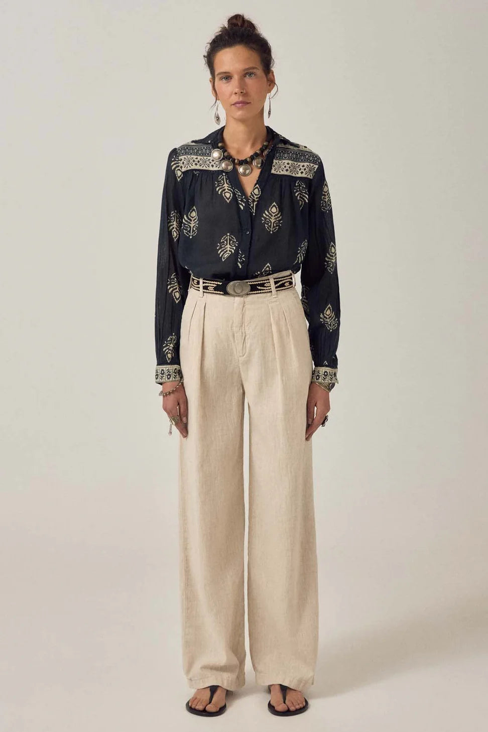 A woman faces forward against a plain background, wearing Maison Hotel's Marina Trousers in sand, a patterned black blouse, black sandals, a belt, and layered necklaces.
