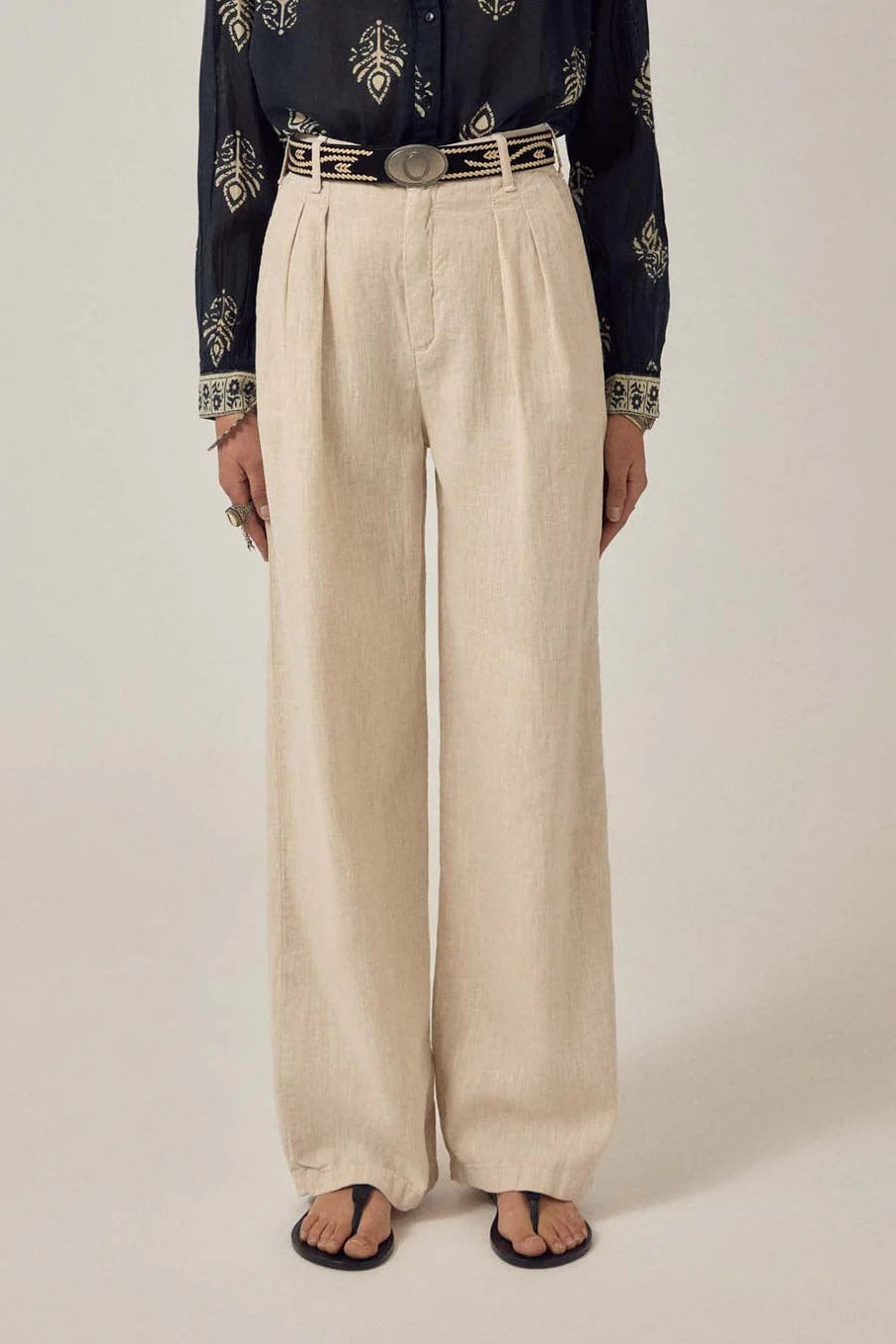 A person wears Maison Hotel’s Marina Trousers in sand, a black patterned shirt, a decorative belt, and black sandals, standing against a plain background.