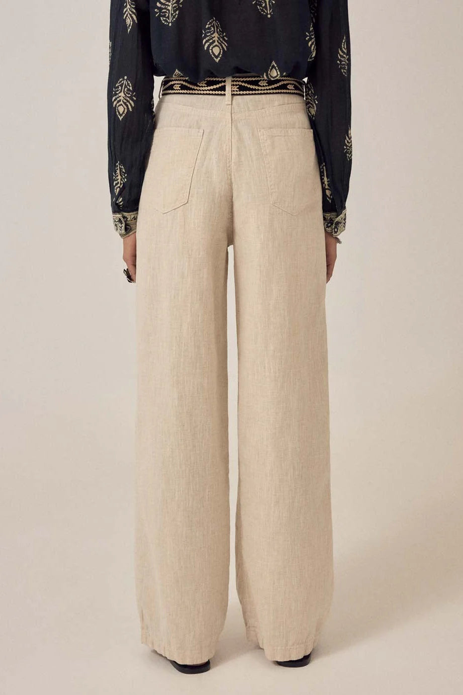 Model wears Maison Hotel's Marina Trousers in Sand, featuring a high-waisted, wide-leg silhouette, paired with a black patterned long-sleeve shirt. Shown from the back against a plain background.