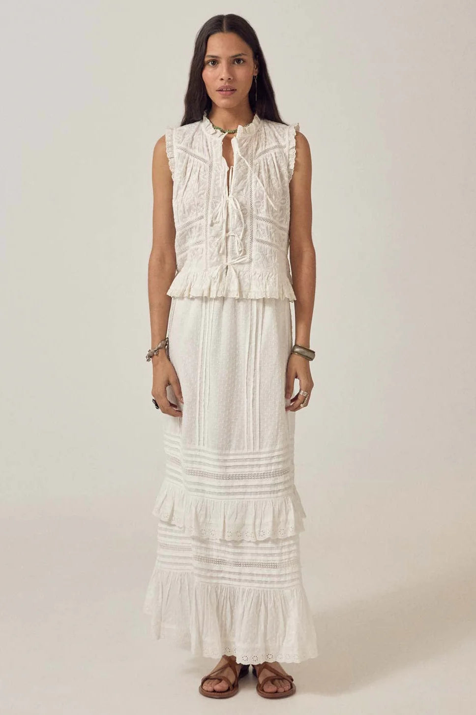 A woman faces forward wearing the Maison Hotel Camila Top in bright white with a long white tiered ruffle skirt and brown sandals, set against a plain background.