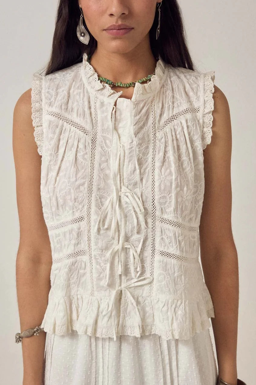 A woman wears the Maison Hotel Camila Top in Bright White, featuring lace trim, ruffled collar, and bow ties, paired elegantly with a matching white skirt.