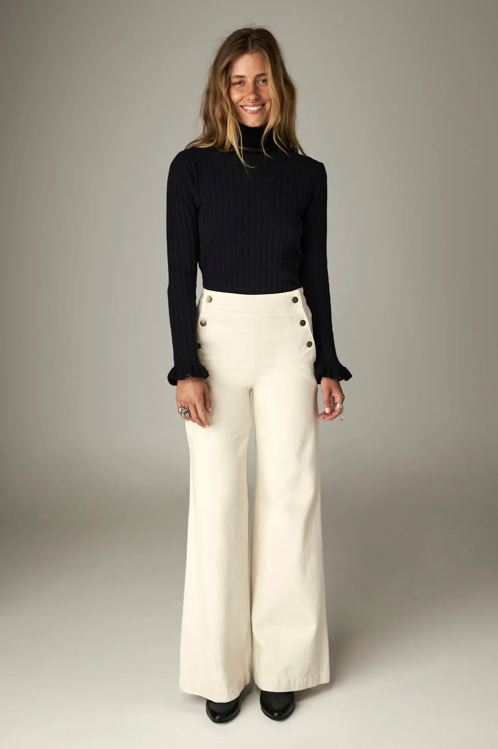 A person wearing the Nancy Sweater - Black Frequency, a black ribbed cable-knit turtleneck with ruffled cuffs, paired with off-white high-waisted wide-leg trousers featuring side buttons.