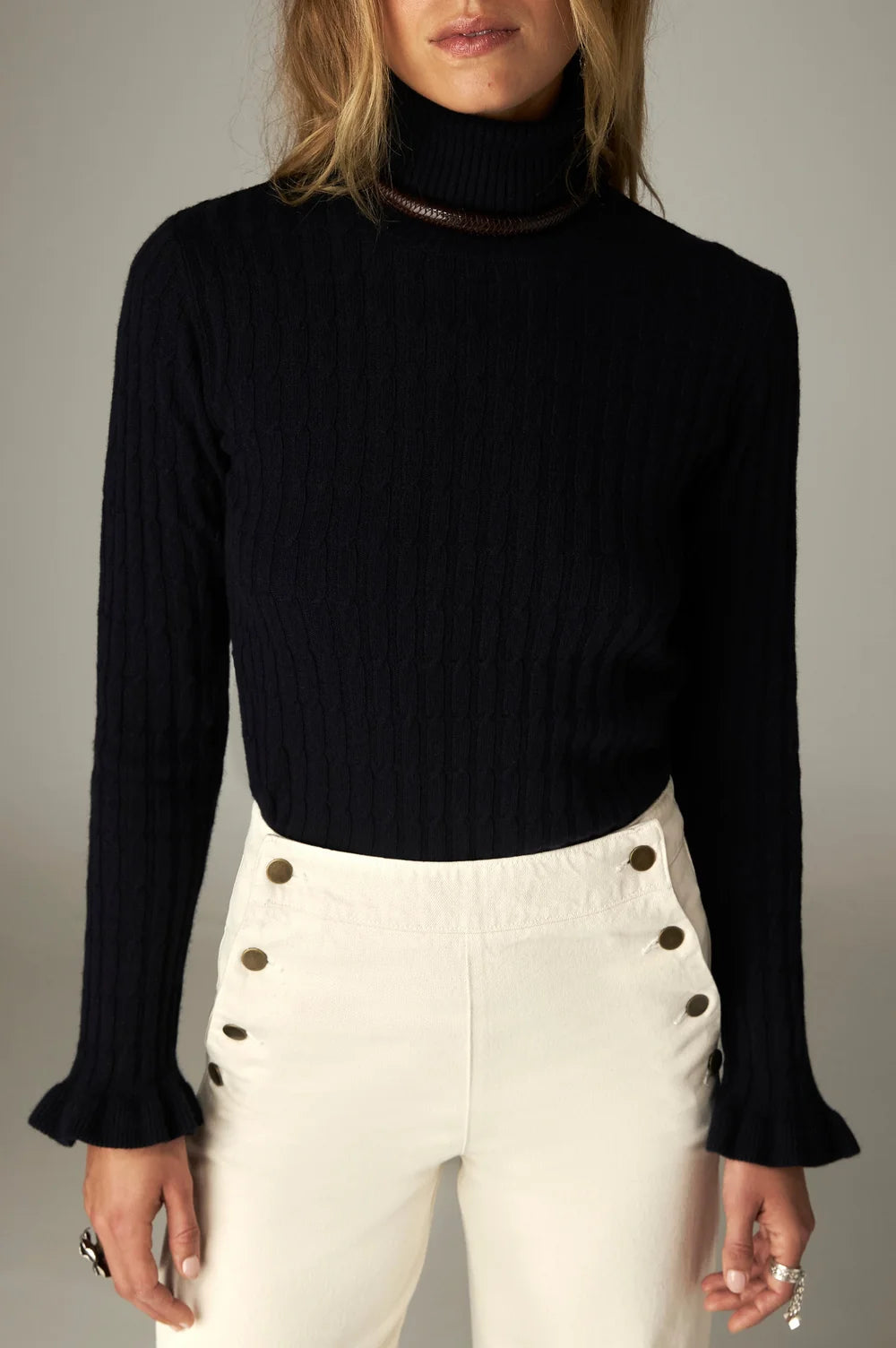 Nancy Sweater - Black Frequency ribbed knit sweater with a mock neckline and ruffled cuffs, worn with white high-waisted pants featuring gold buttons.