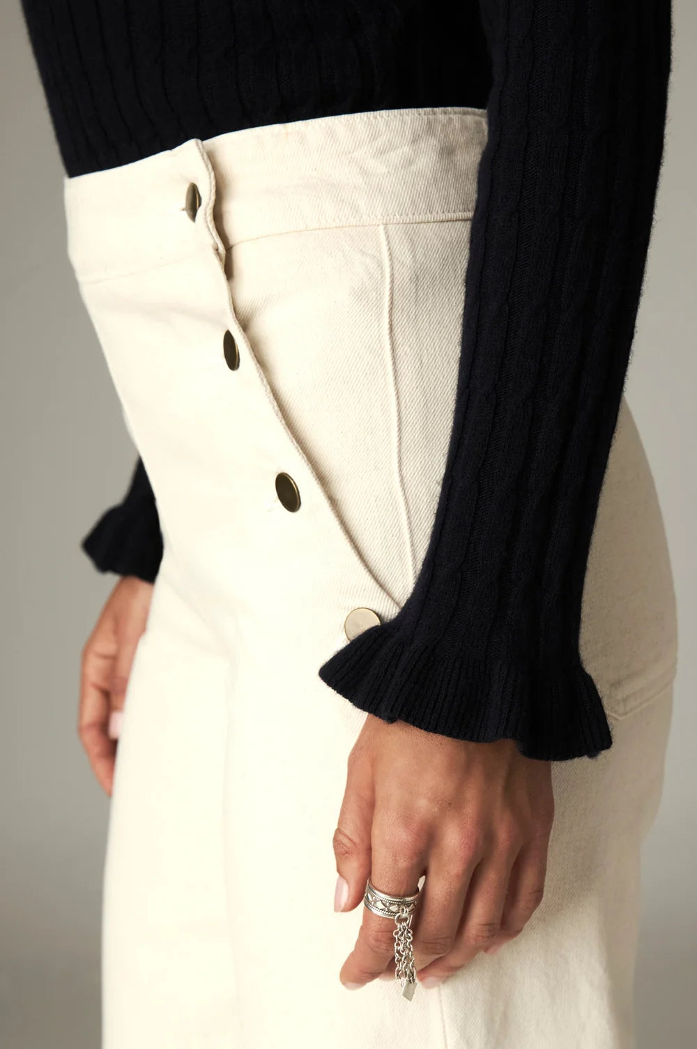 Nancy Sweater - Black Frequency ribbed knit sweater with a mock neckline and ruffled cuffs, tucked into high-waisted off-white trousers with a side button closure.