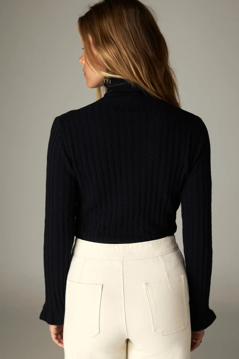 Back view of a person wearing a black Nancy Sweater - Black Frequency with a cable-knit mock neckline and ruffled cuffs, paired with white pants.