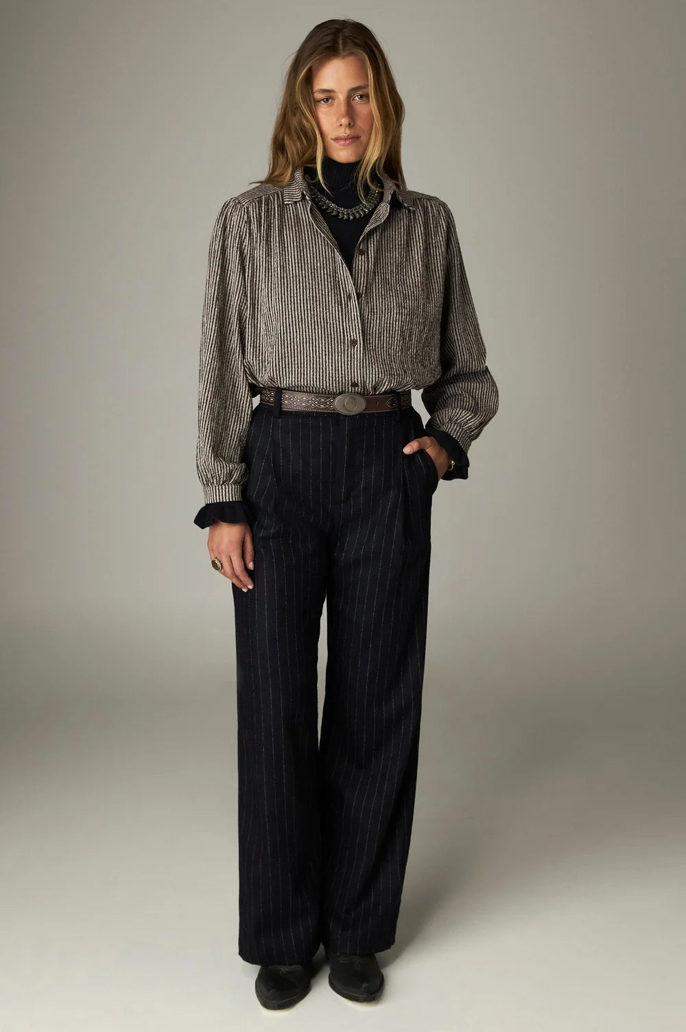A Maison Hotel Claude Blouse - Coffee Stripes with a gathered yoke and dark vertical stripes worn over a black turtleneck, paired with navy pinstriped trousers and a brown leather belt.