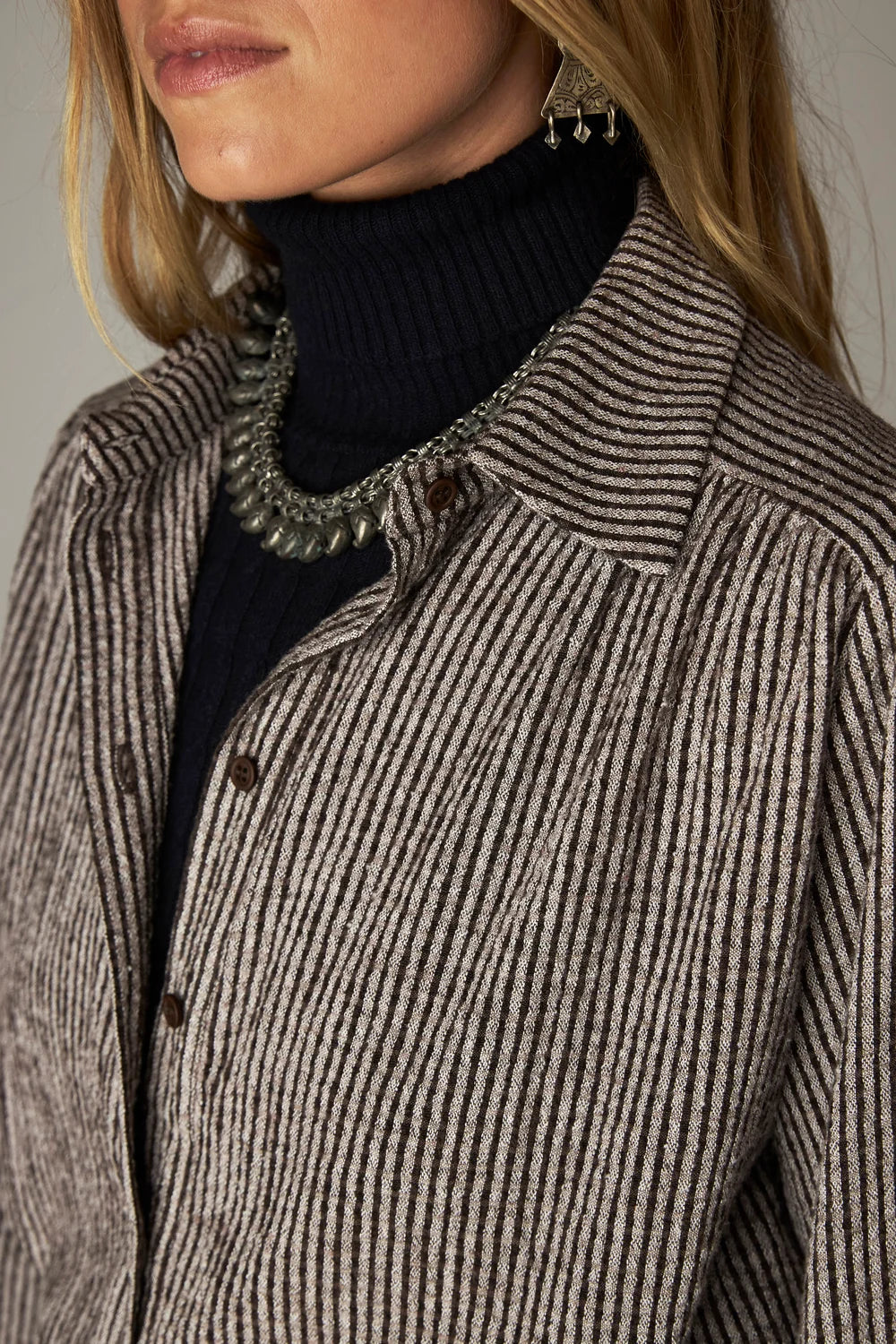 A woman wears a coffee-striped Maison Hotel Claude Blouse - Coffee Stripes over a black turtleneck, accessorized with a metallic collar necklace.