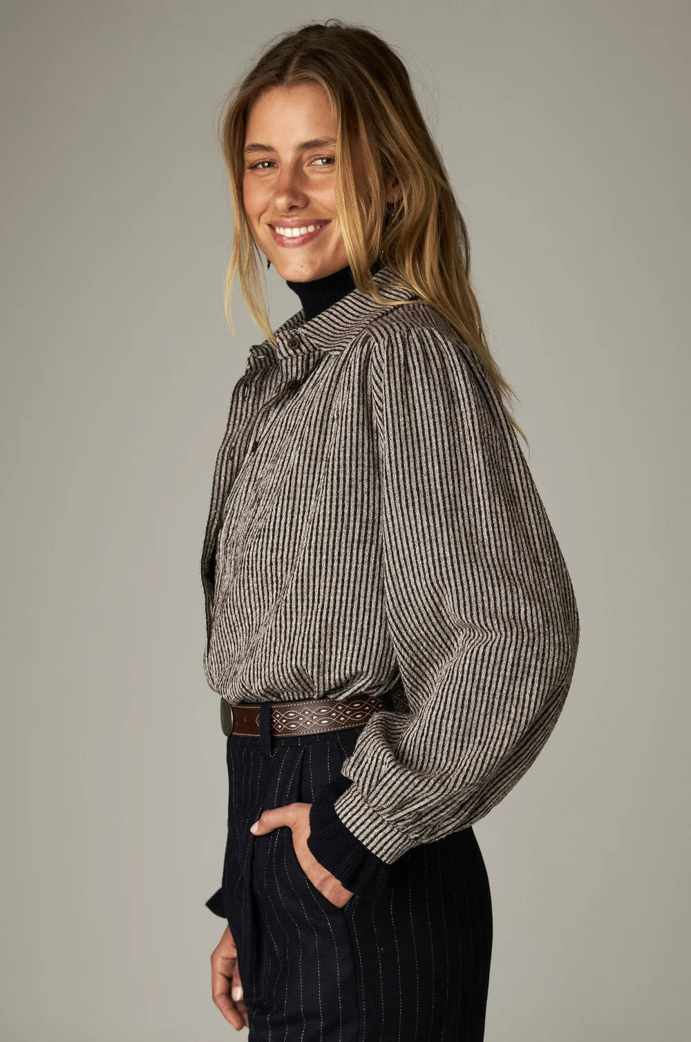 A person wears a Maison Hotel Claude Blouse - Coffee Stripes gathered yoke blouse over a dark turtleneck, paired with dark pinstriped trousers and a patterned leather belt.