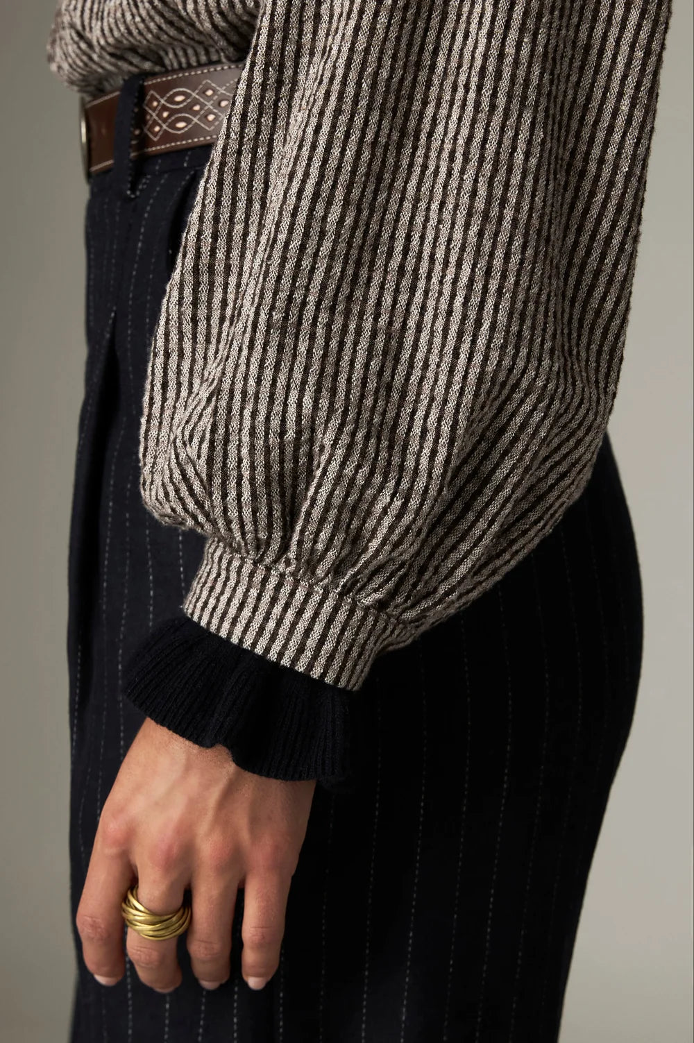 A Maison Hotel Claude Blouse - Coffee Stripes with gathered puff sleeves and black ribbed cuffs, worn tucked into dark pinstriped trousers with a brown belt.