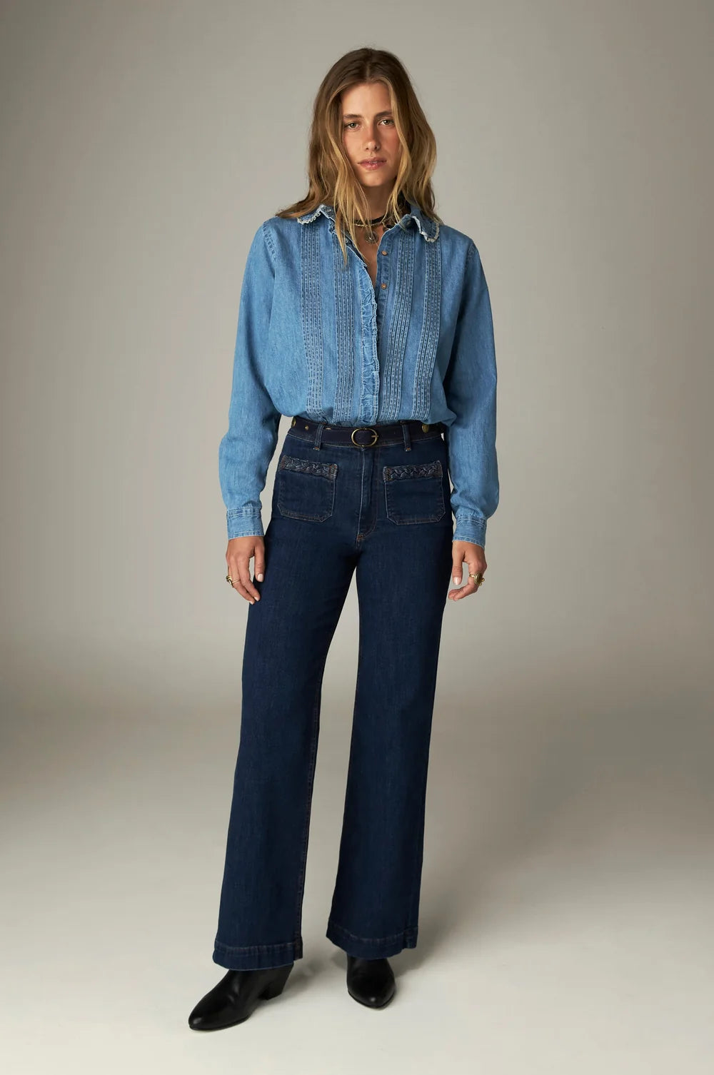 A woman wears a long-sleeved denim shirt tucked into Giulietta Pant - Blue Denim, featuring a high-waisted, wide-leg cut with front patch pockets and a black belt.
