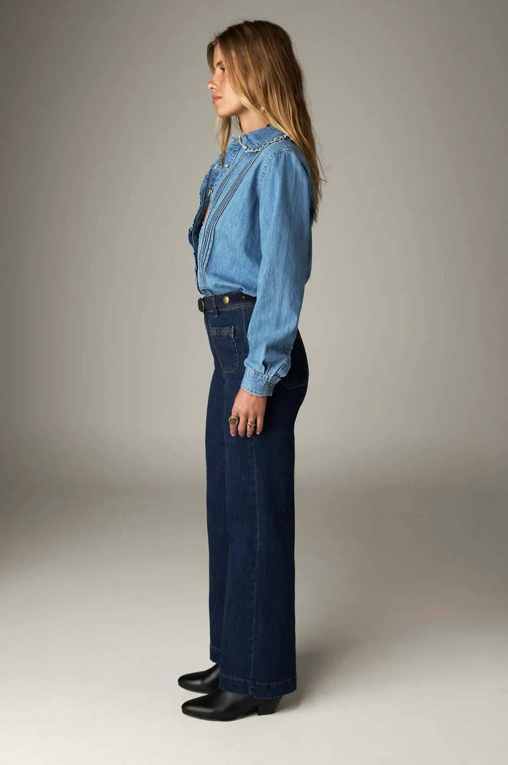 A person wearing a long-sleeved light denim shirt with a ruffled collar and Giulietta Pant - Blue Denim high-rise wide-leg pants.