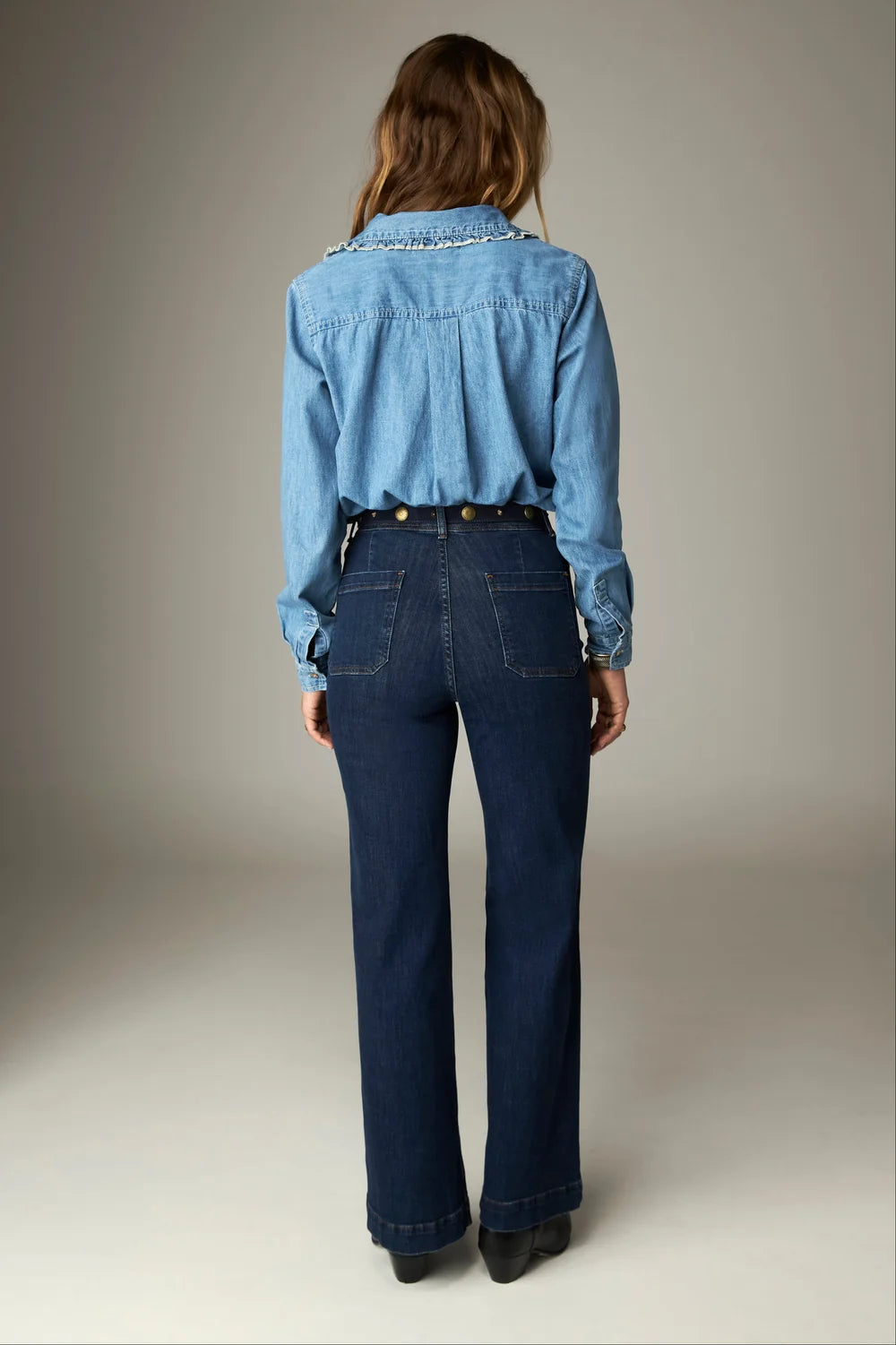 Back view of a person wearing a denim shirt with a ruffled collar tucked into Giulietta Pant - Blue Denim, featuring high-waisted, dark-wash blue jeans with back patch pockets and metal waist buttons.