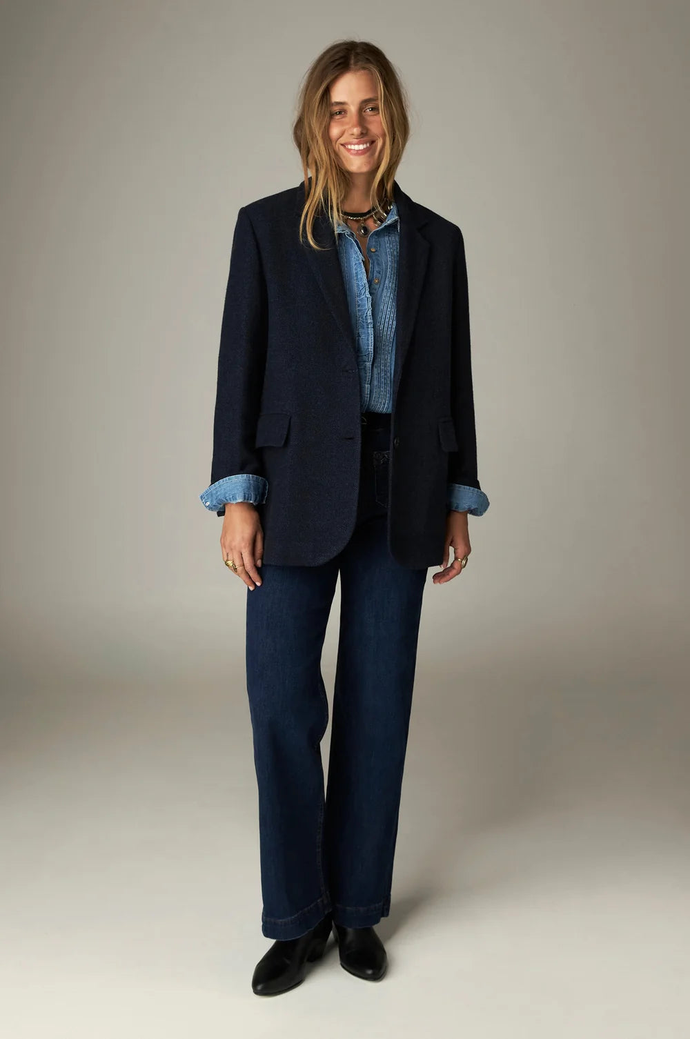 A person wearing a dark blazer over a denim shirt, Maison Hotel Giulietta Pant - Blue Denim high-rise wide-leg pants, and black boots.