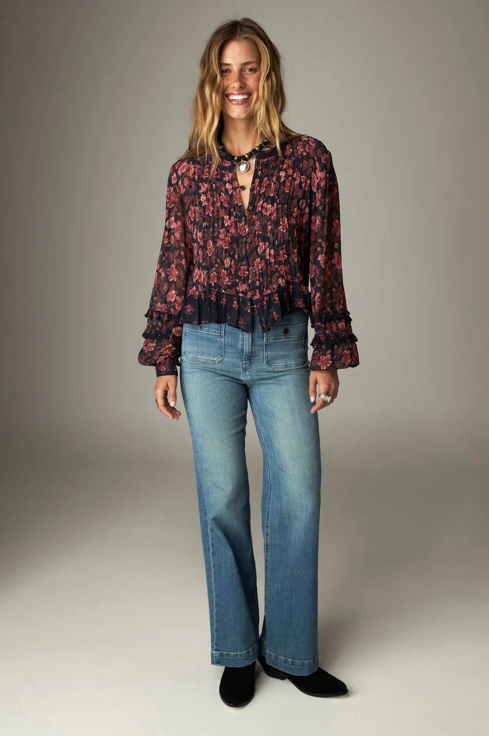 Woman wearing a dark floral Lerele Blouse - Punk Pink with a ruffled hem, tucked into blue wide-leg jeans.