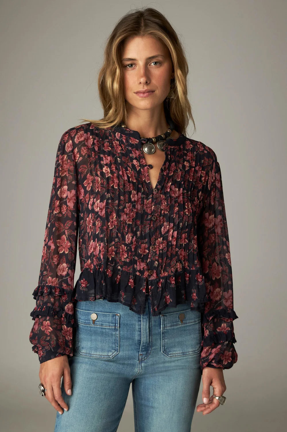 A woman wears a bohemian Maison Hotel Lerele Blouse - Punk Pink featuring pleated chest detailing, a ruffled hem, and ruffled cuffs, paired with blue jeans.
