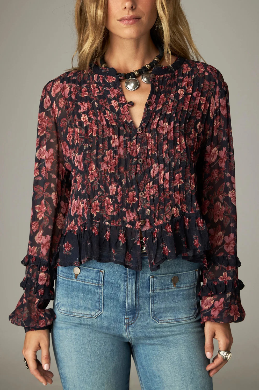 A woman wears a Maison Hotel Lerele Blouse - Punk Pink featuring a bohemian, long-sleeved navy-blue design with a pink floral pattern, pleated front, and ruffled cuffs, paired with blue jeans.