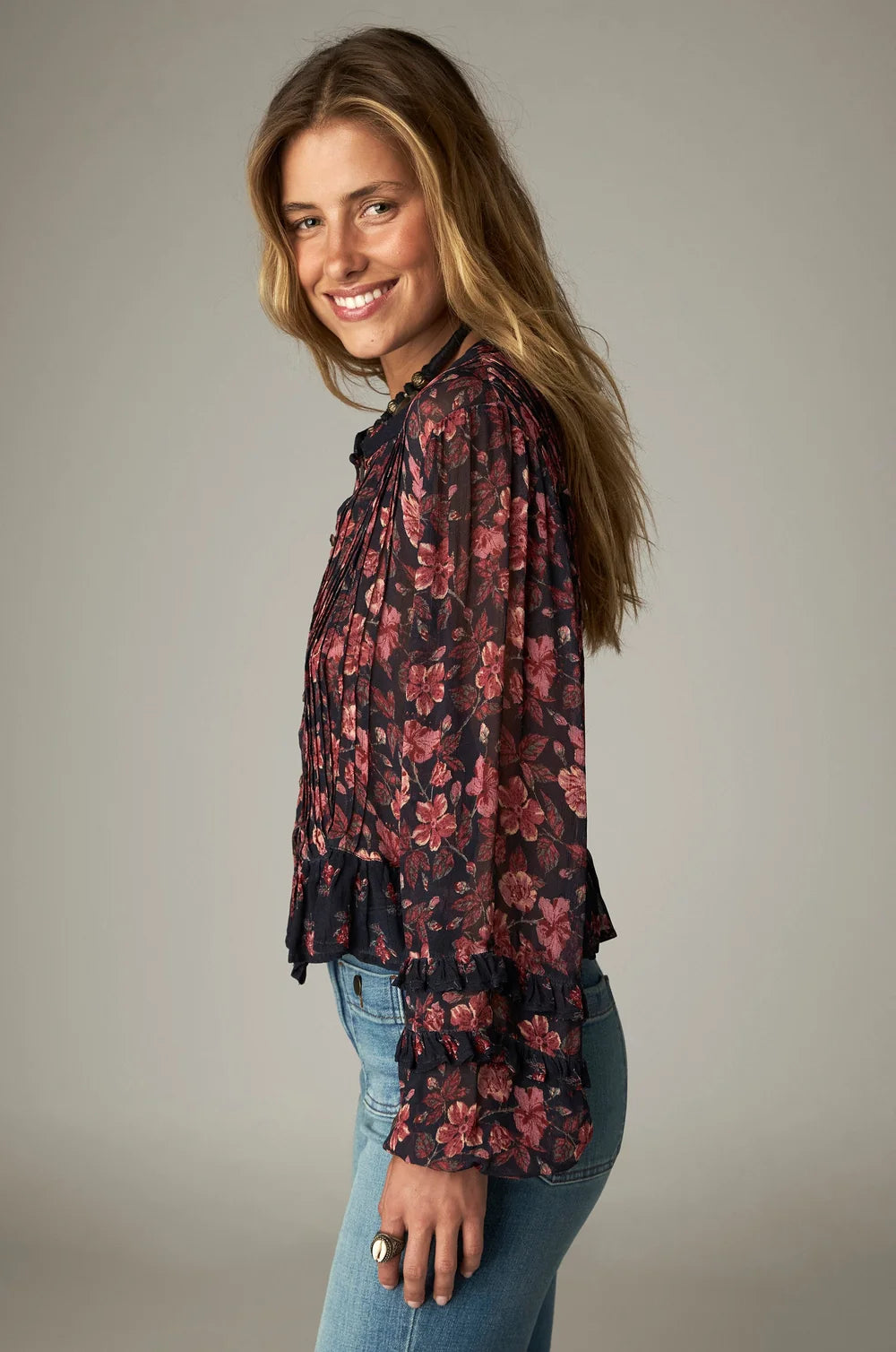 A woman smiles in a dark pink floral-print Maison Hotel Lerele Blouse - Punk Pink with a relaxed silhouette, pleated detailing, and ruffled cuffs, paired with blue jeans.