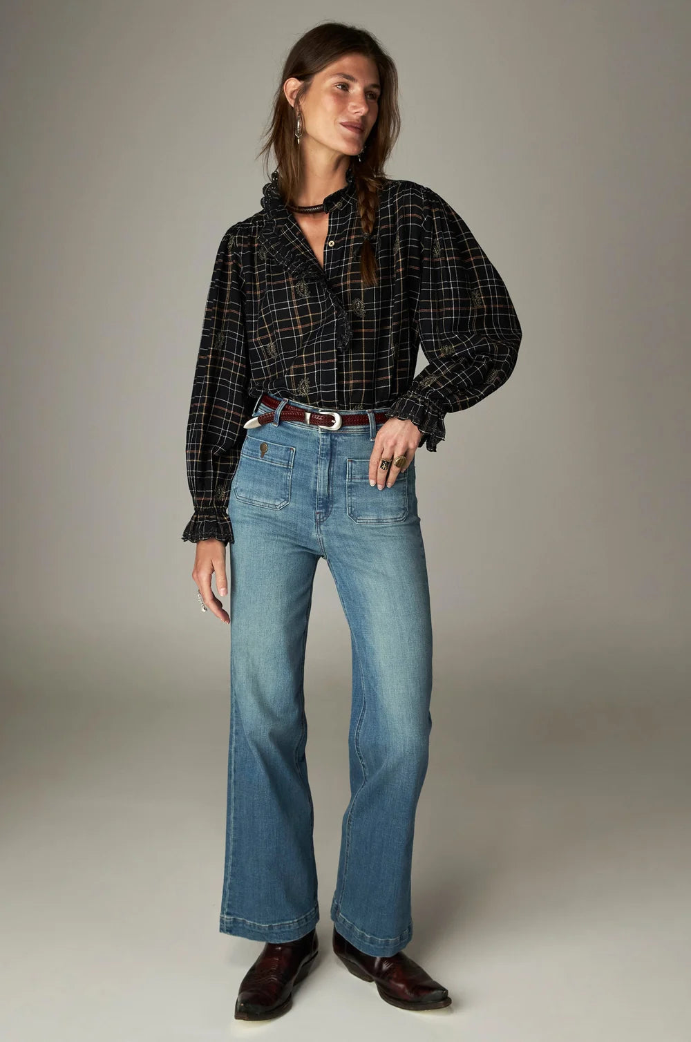 A woman wears a long-sleeved, ruffled Maison Hotel Coco Blouse - Noir Plaid tucked into light-wash blue jeans with a brown leather belt and dark boots.
