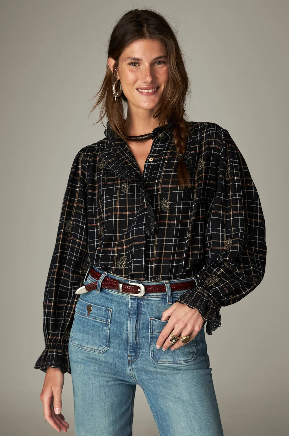 A woman wearing a long-sleeved Maison Hotel Coco Blouse - Noir Plaid with a ruffled collar and cuffs tucked into light-wash denim jeans with a brown leather belt.