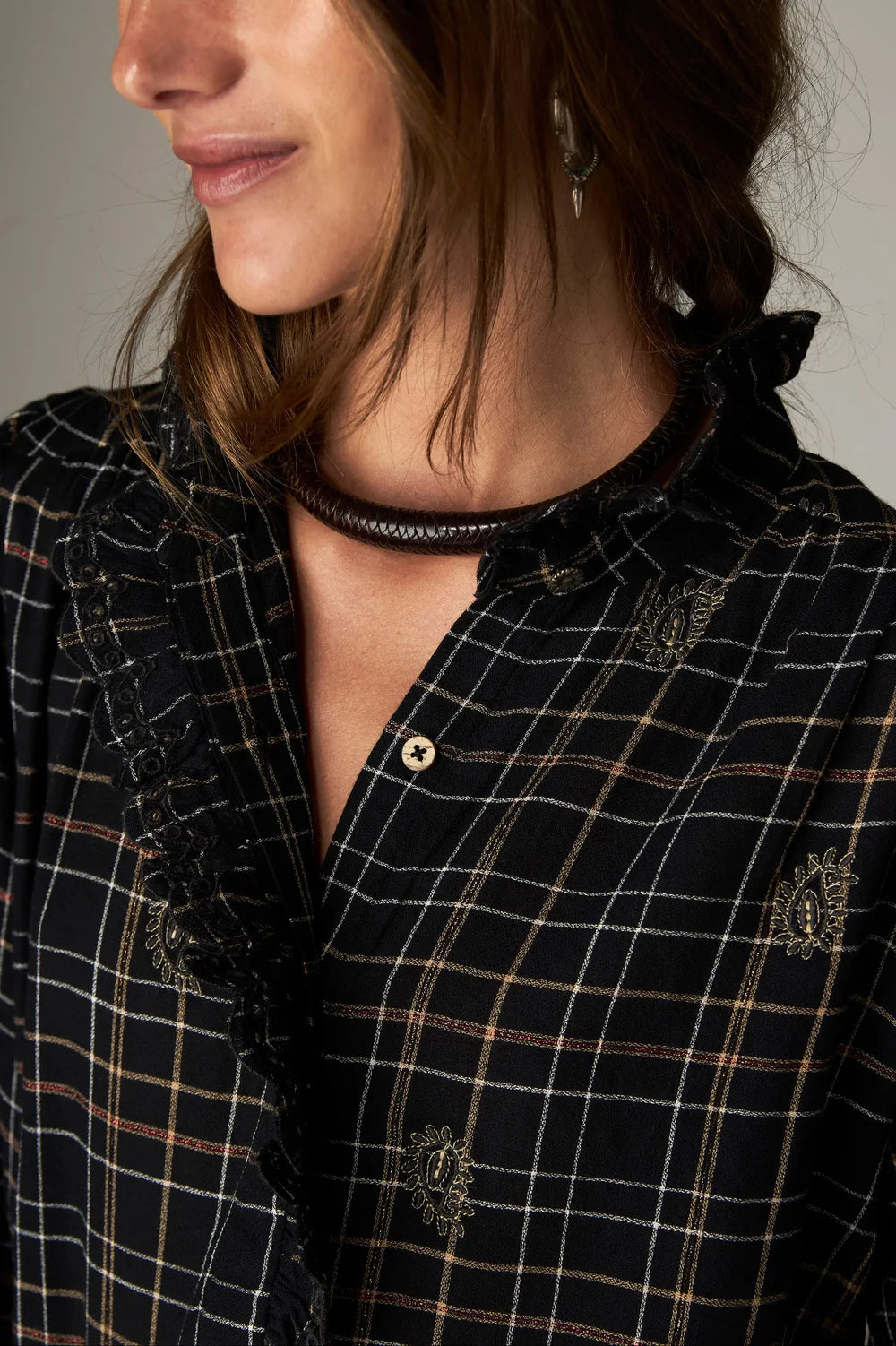 Close-up of a person wearing a Maison Hotel Coco Blouse - Noir Plaid, featuring ruffles along the placket and small embroidered floral details.